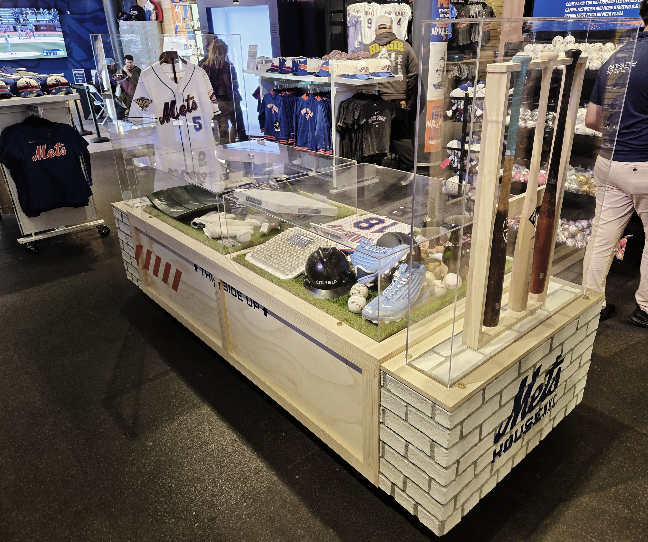 A retail display case shaped like a baseball field on a wooden stand, showcasing baseball memorabilia such as baseballs, bat, glove, and jersey, with a brick pattern design on the sides. The store sells New York Mets merchandise and is located near C