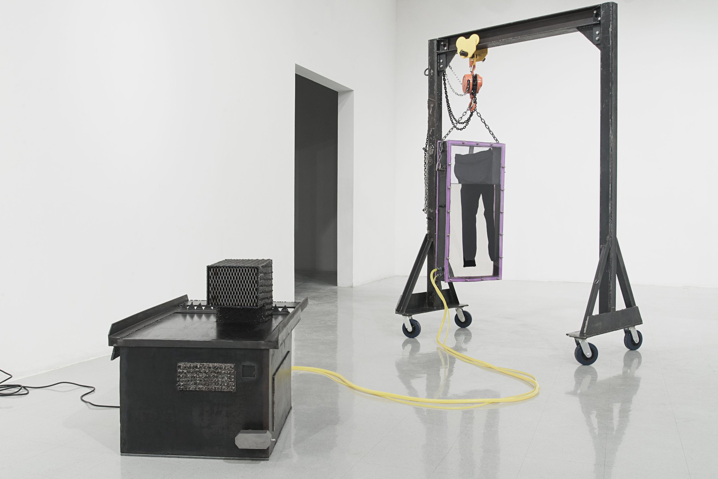 Art installation featuring a black metal frame with chains, a framed pair of black pants, and various climbing and industrial equipment in a white gallery space.