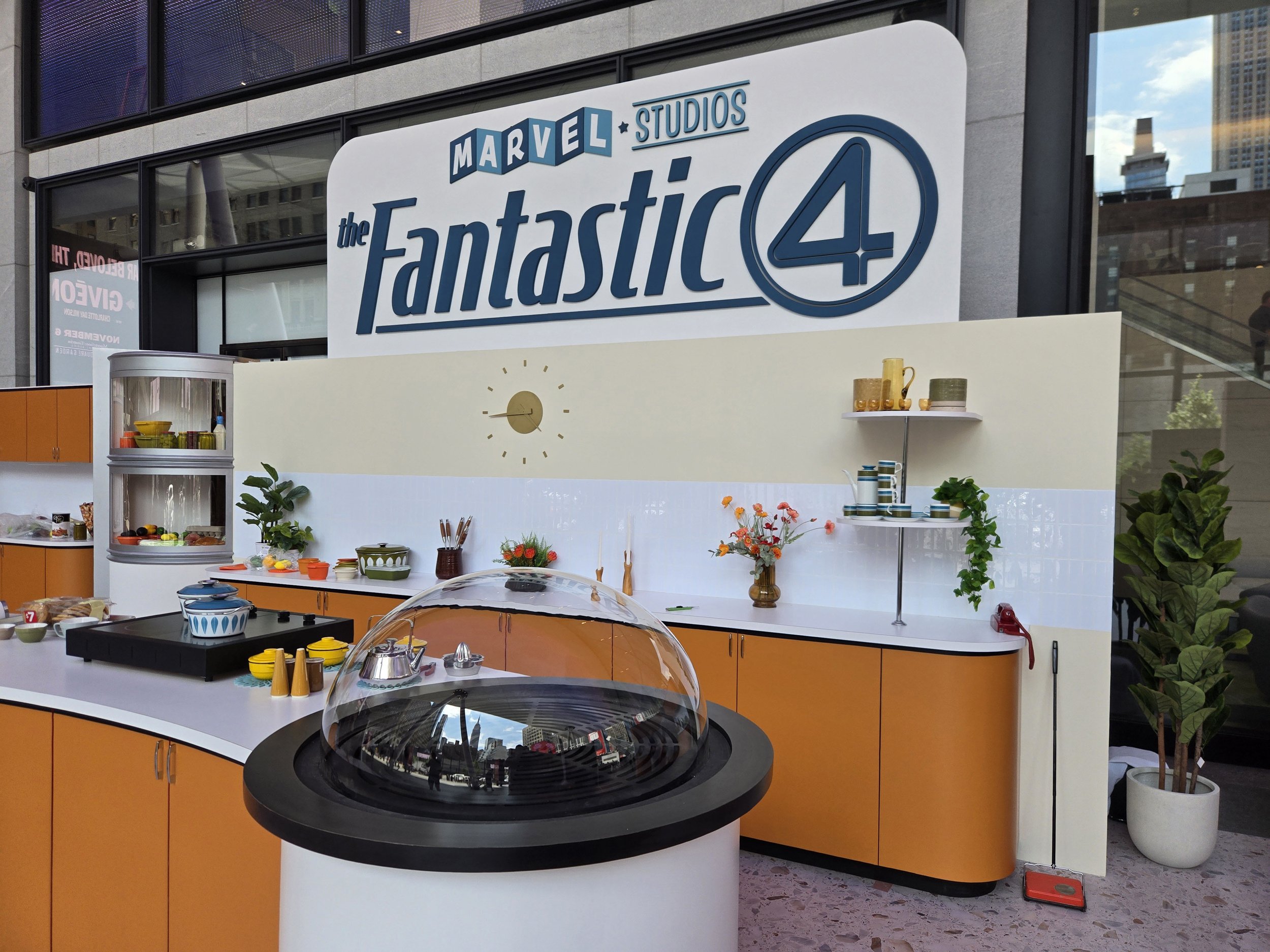 An outdoor food display stands in front of a high-rise building, with a sign reading "Marvel Studios The Fantastic Four." The display features various bowls and containers of food and colorful flowers, alongside a black round hot plate and a clear co