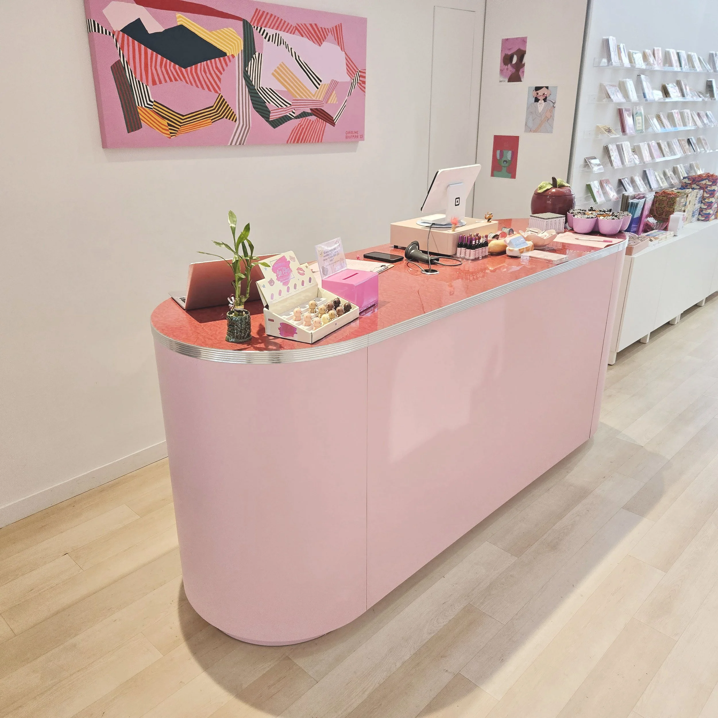 A pink counter with small figurines, a plant, and various pink and colorful items on display. Wall art includes a large abstract pink painting and smaller illustrations, with a white shelf holding books or pamphlets.