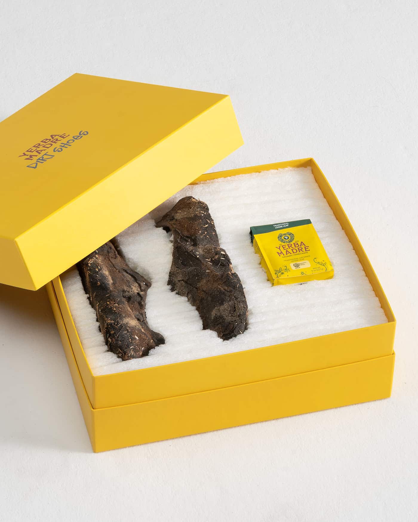 A yellow box with the words "Yerba Mate Art Shoes" on the lid, containing two dried yerba mate roots and a yellow packet of yerba mate.