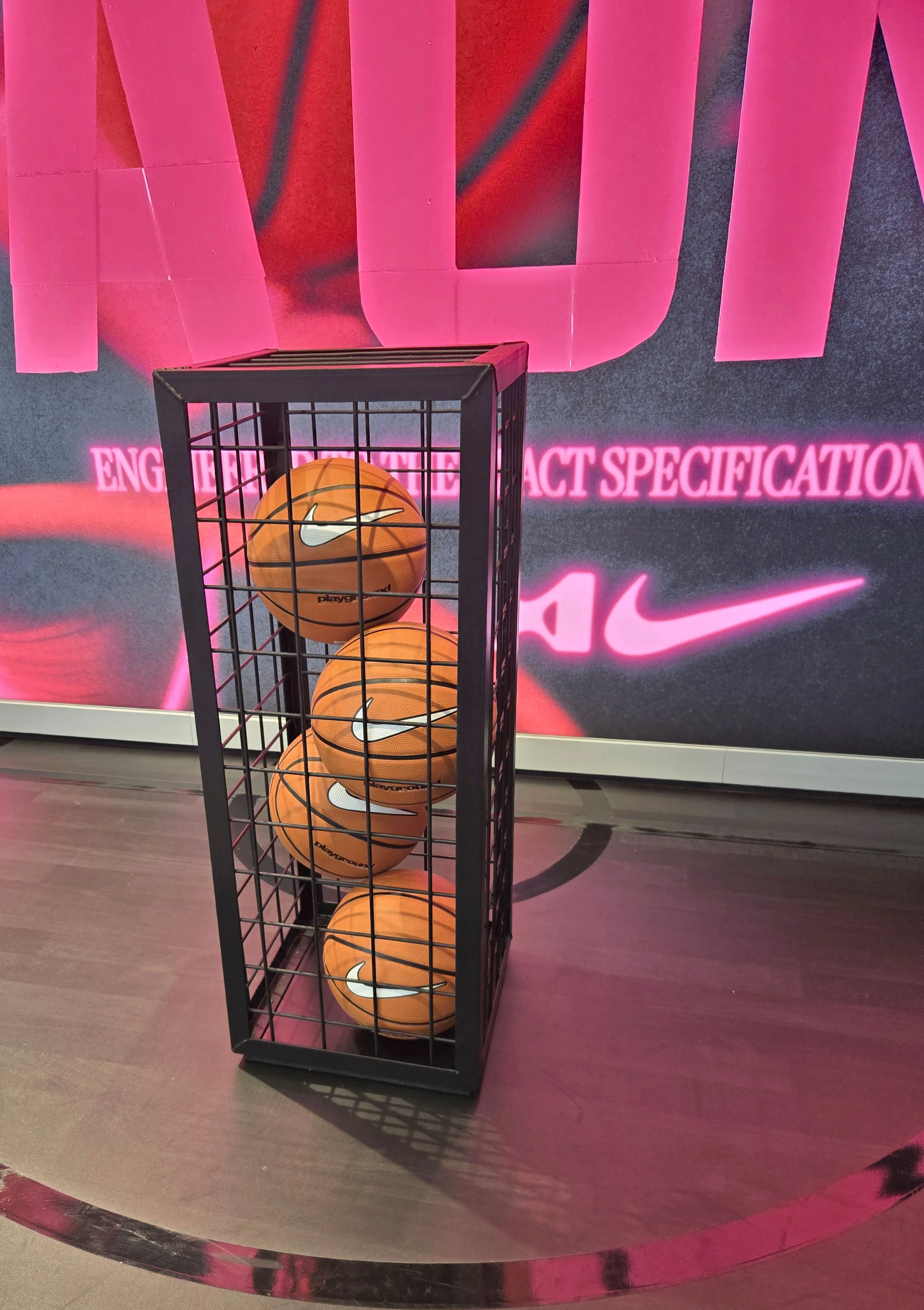 Basketball court with a black metal rack holding orange Nike basketballs in front of a pink Neon sign that reads 'ENGAGEMENT IMPACT SPECIFICATION' and a pink Nike logo.