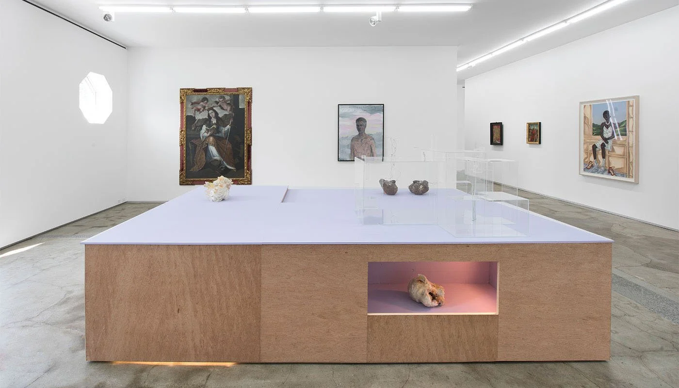 Interior of an art gallery with various paintings on white walls, a wooden display table with sculptures, and a concrete floor.