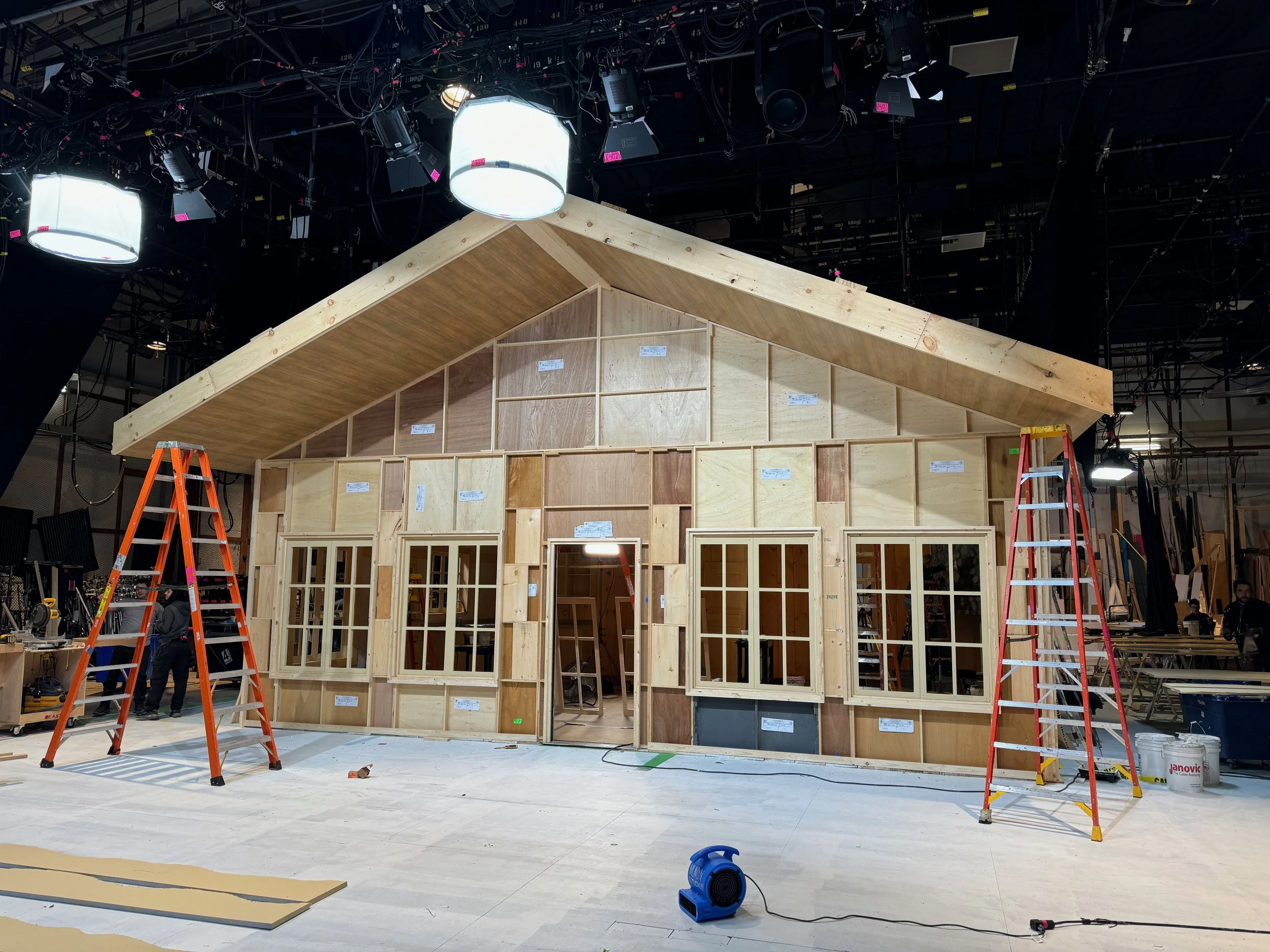 A stage under construction with wooden framework, ladders, and construction equipment, in an indoor setting with stage lighting and overhead cables.
