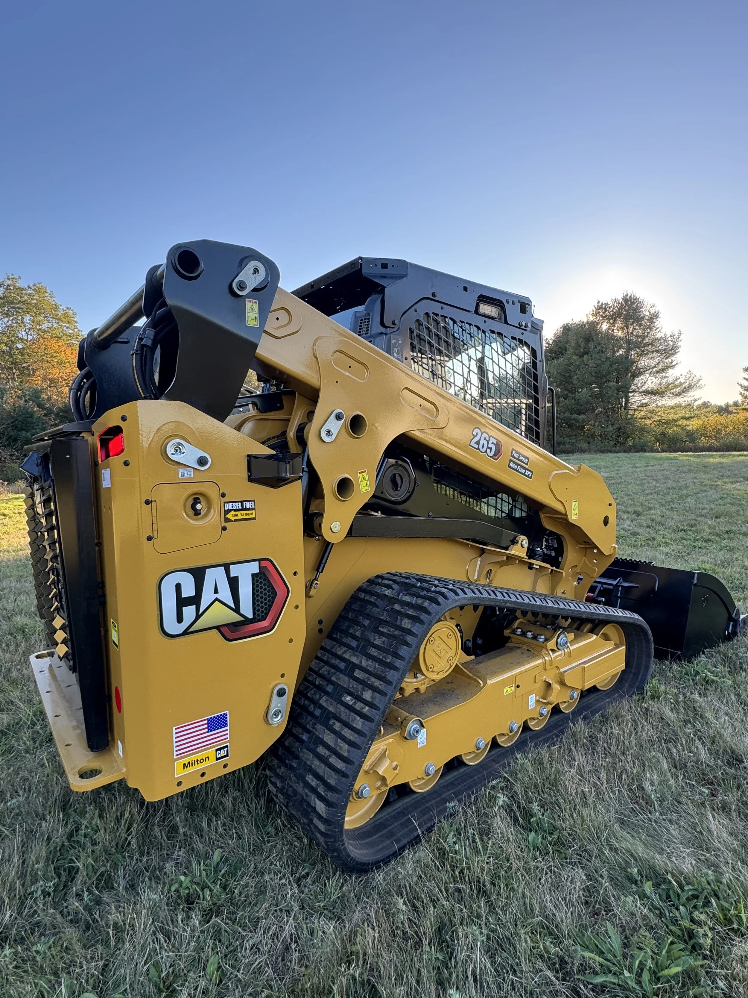 A yellow CAT compact track loader with black tracks parked on a grassy field during sunset, with trees in the background.