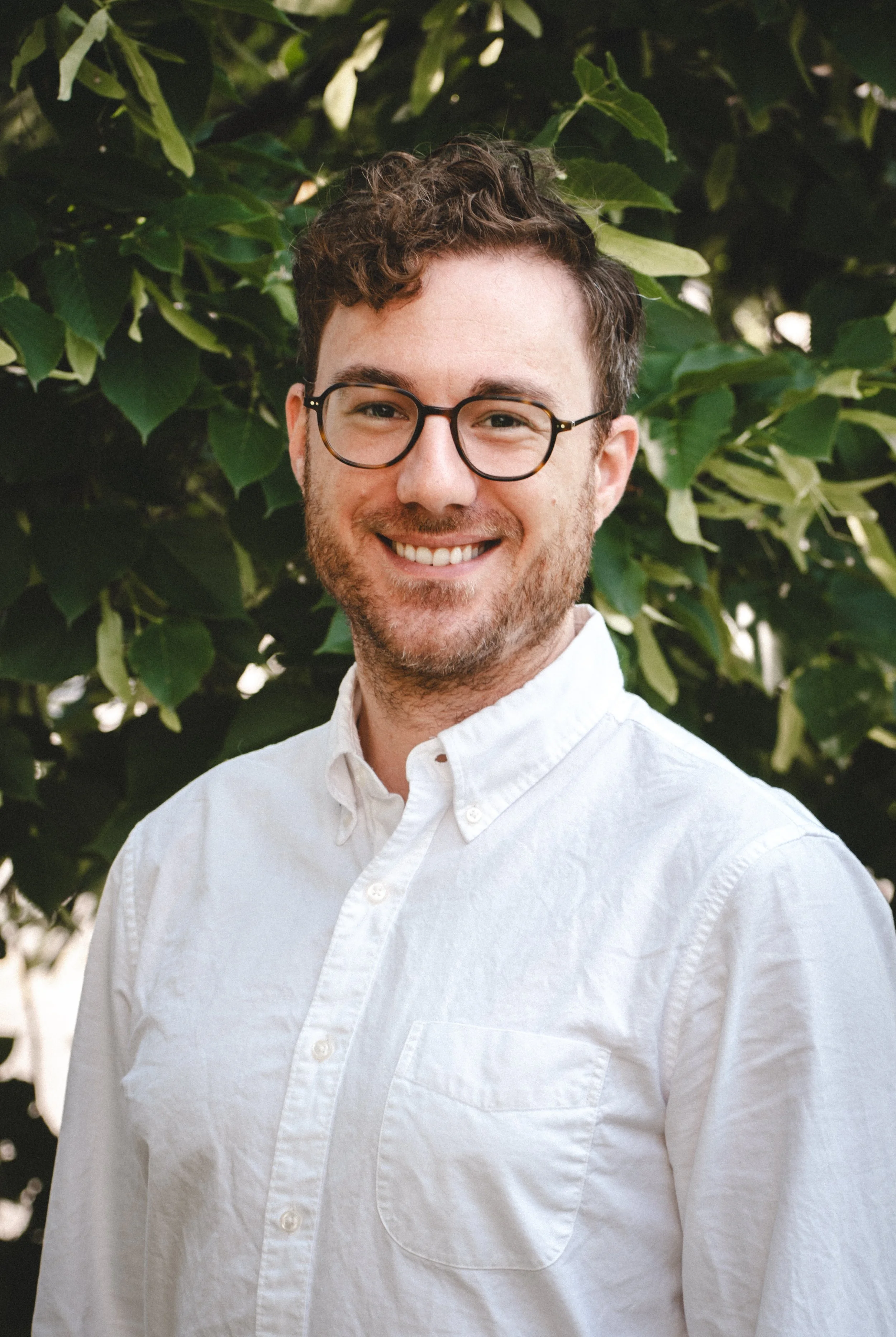 A young man professional therapist with brown curly hair, glasses, and a warm kind smile. He appears welcoming and thoughtful.