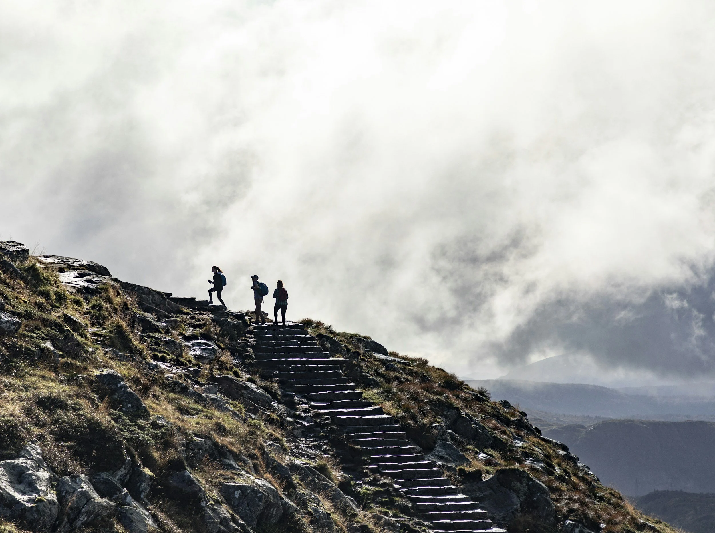 A group of four hikers ascending a mountain trail on rocky steps, with a cloudy sky overhead and rugged terrain in the background.