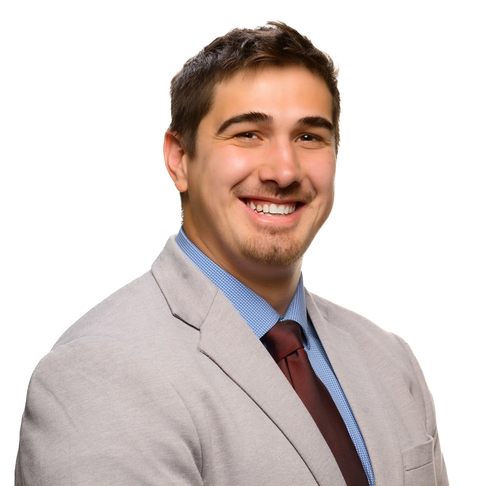 Close-up photo of a smiling young man with dark hair, wearing a light gray suit, blue dress shirt, and a brown tie, against a white background.