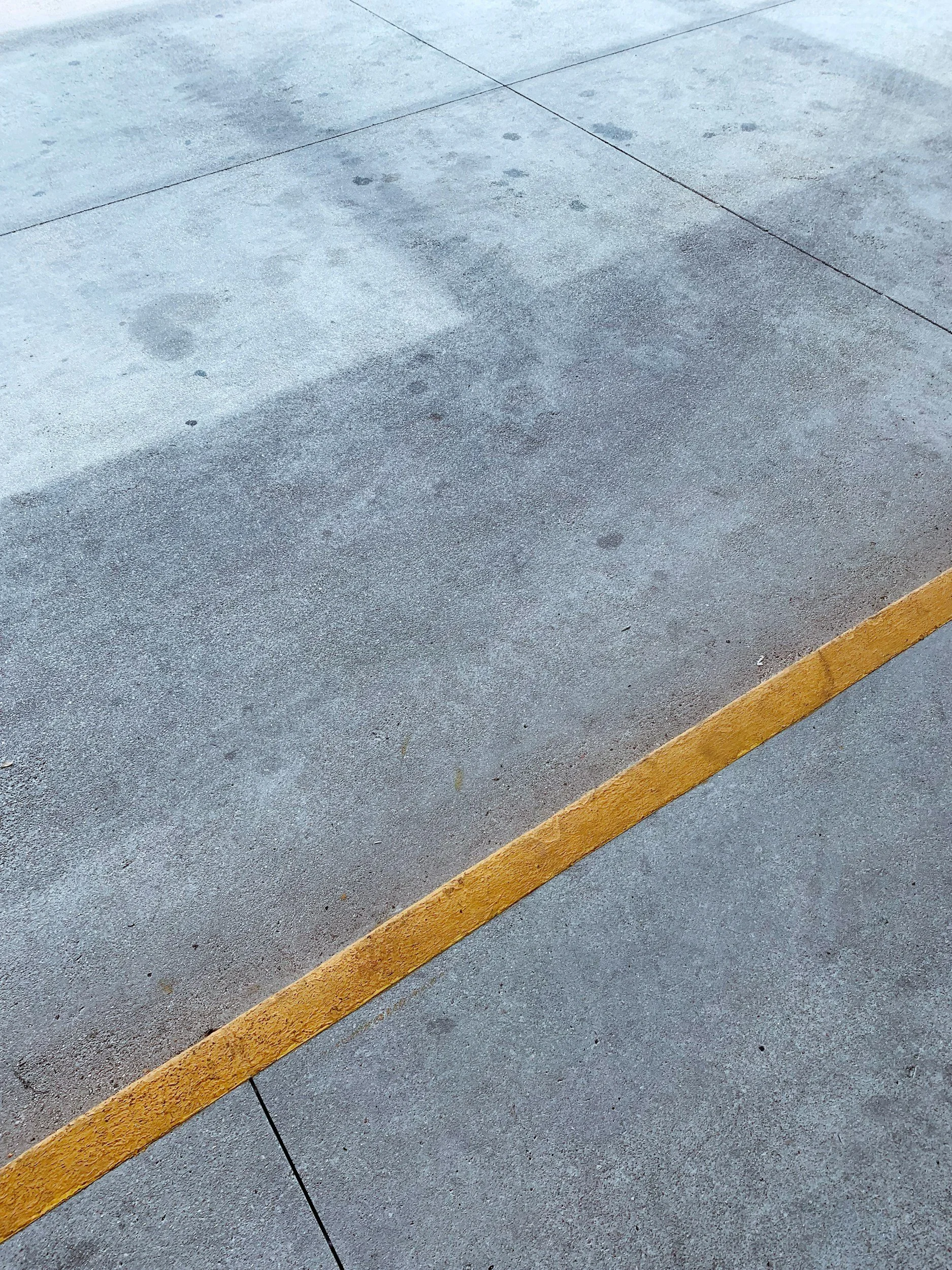 A concrete sidewalk with a yellow painted line along the bottom edge, showing some surface imperfections and small dark spots.