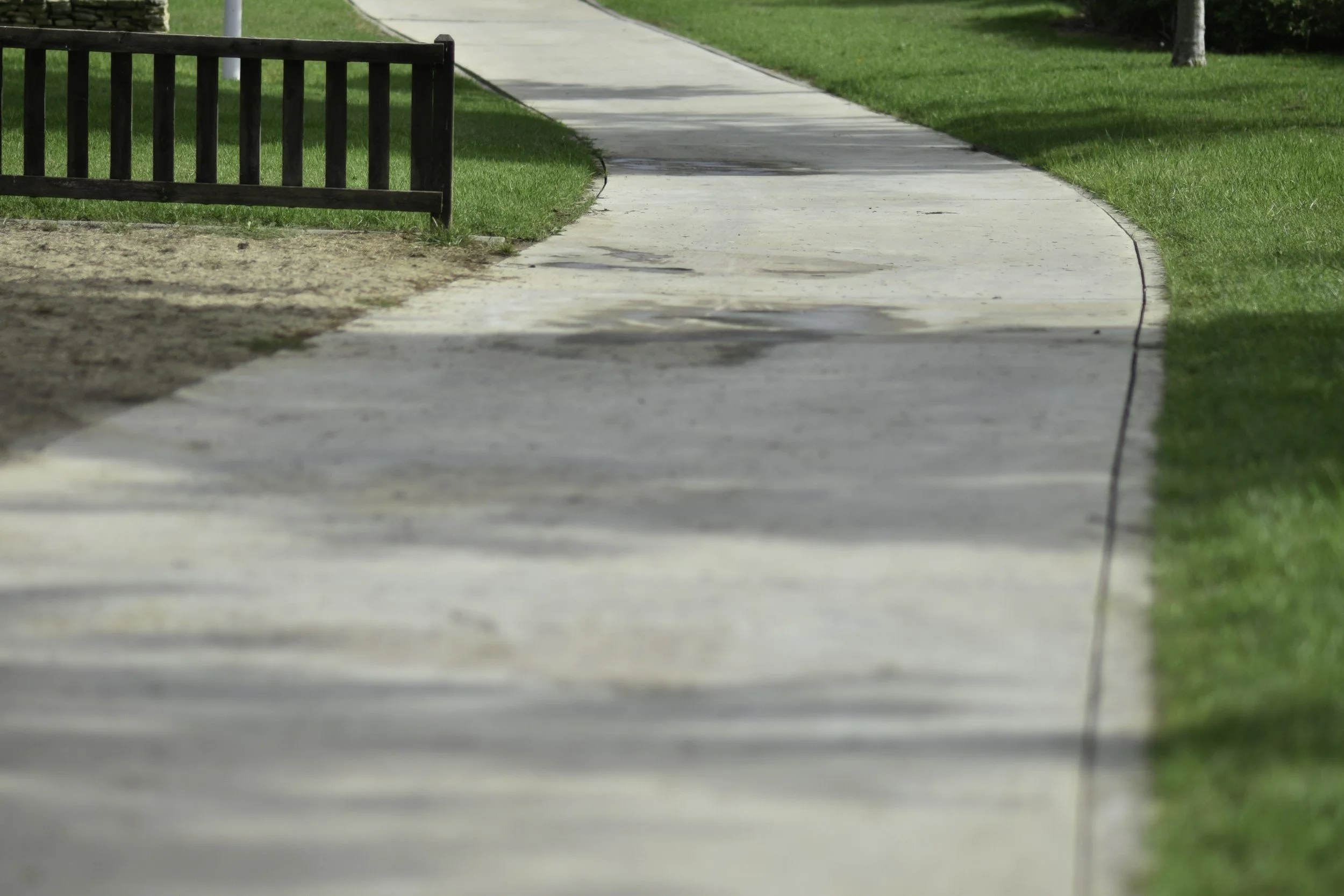 A sidewalk in a park with green grass on both sides and a small black fence on the left side.