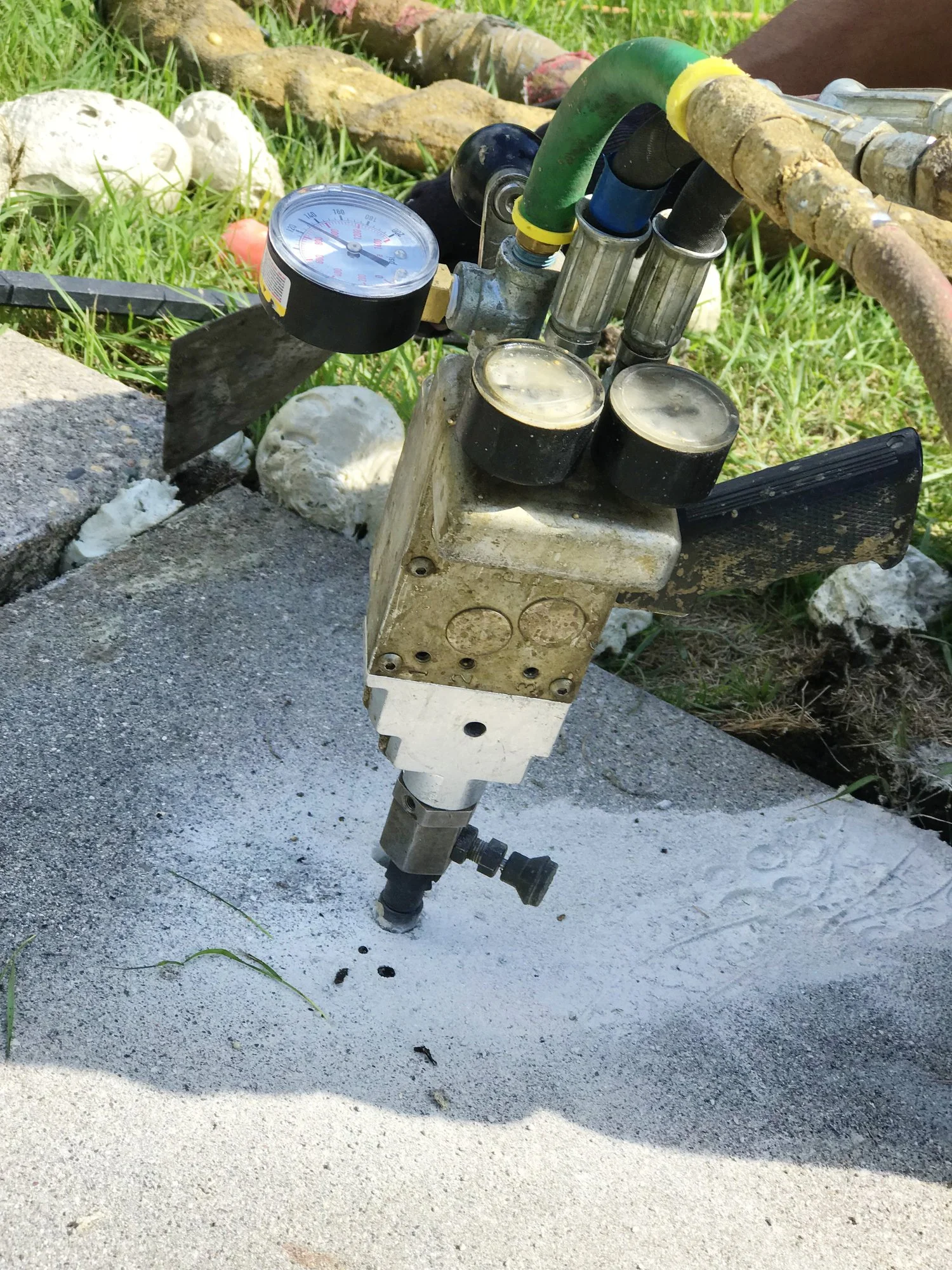 This is a photograph of a piece of construction or industrial equipment installed on a concrete surface. The equipment has a pressure gauge, several pipes, and black caps or controls. The surrounding area includes grass, rocks, and muddy areas.