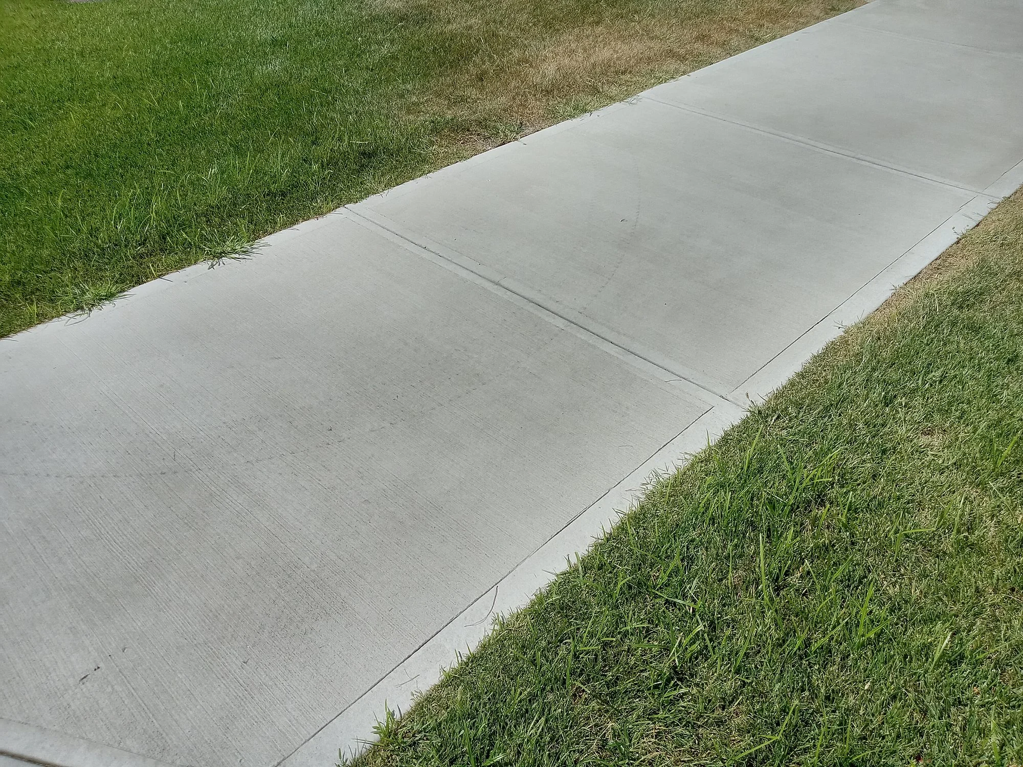 A flat and even concrete sidewalk running alongside a grassy lawn on a sunny day.