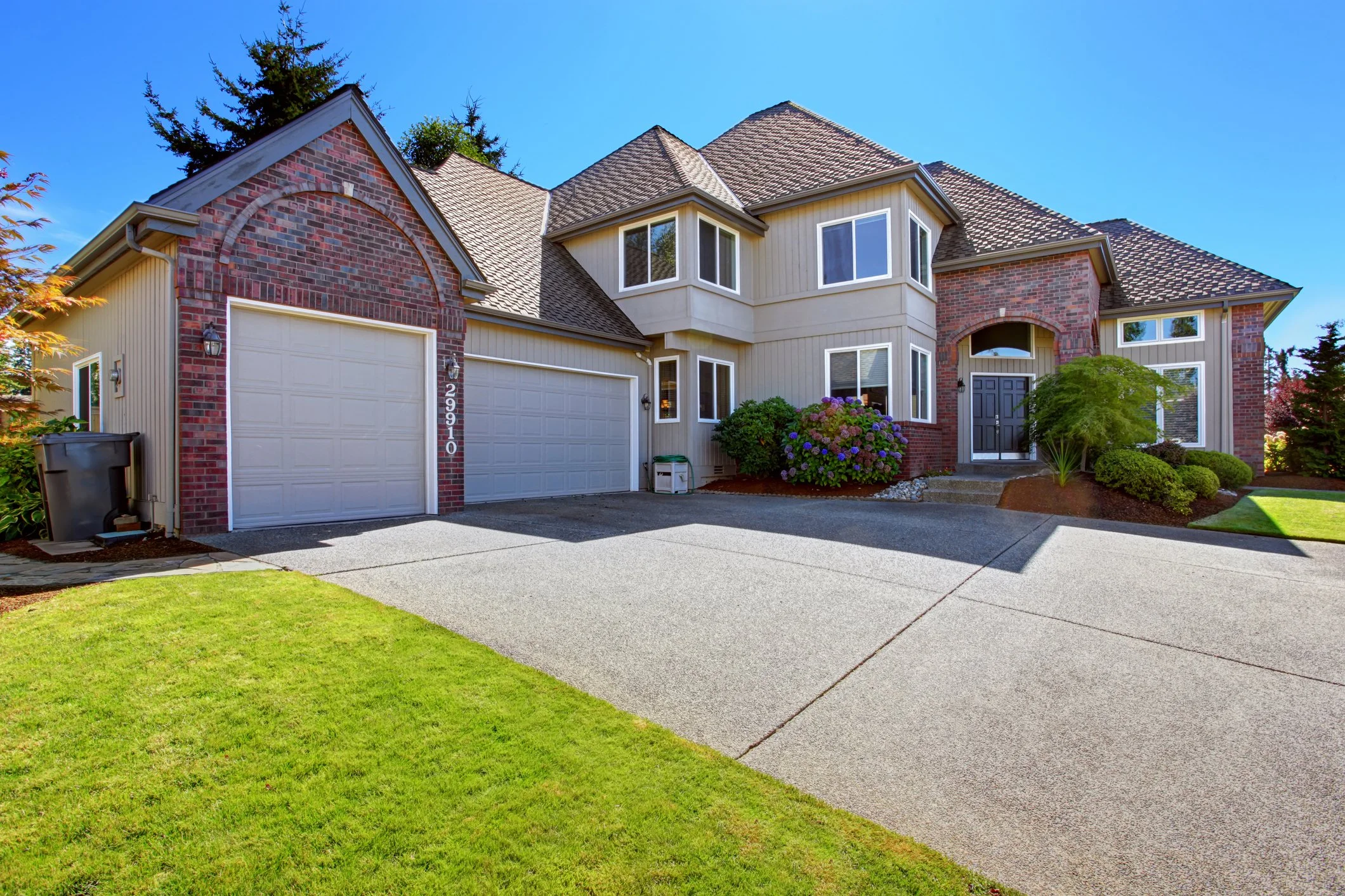 A two-story house with a brick facade, beige siding, and a gabled roof, featuring a double garage door, surrounded by a lawn, shrubs, and trees under a clear blue sky.
