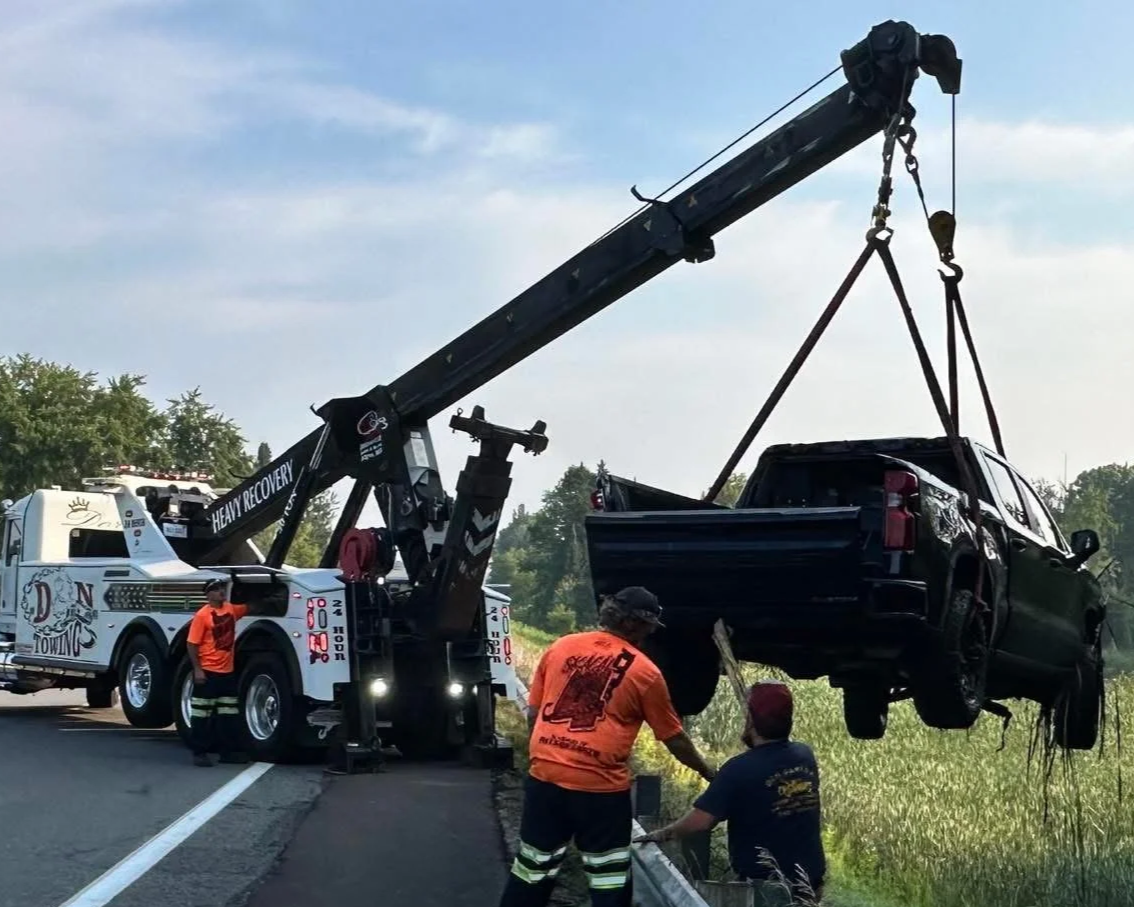 Tow truck lifting a black SUV onto the roadside. Two workers are assisting, one in an orange shirt and the other in a blue shirt, with in a grassy area beside the road.