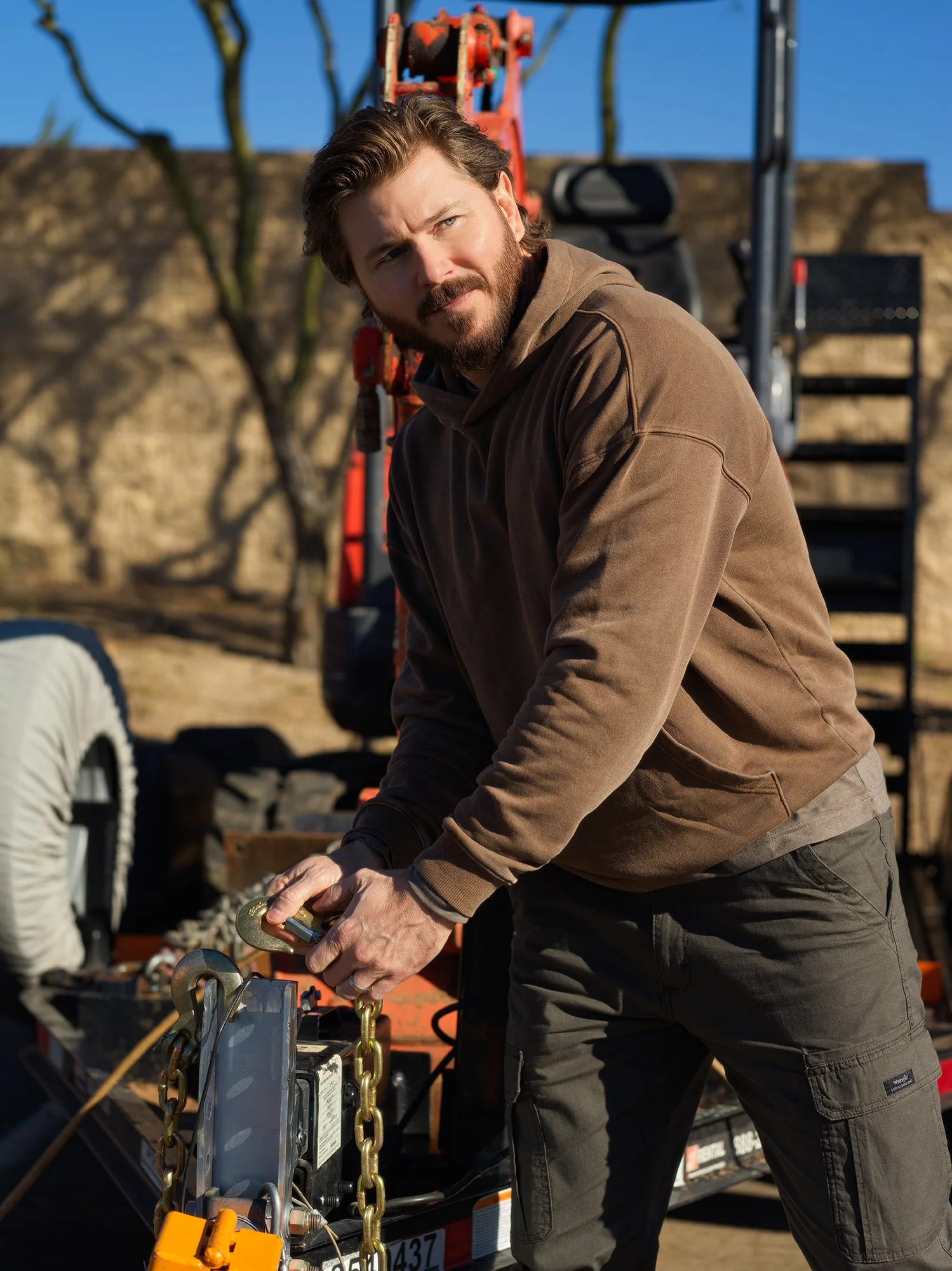 A man with brown hair and a beard, wearing a brown hoodie and dark pants, is working with electrical equipment outdoors during daytime. He appears to be connecting or inspecting the device on a trailer, with tools and chains visible. In the backgroun