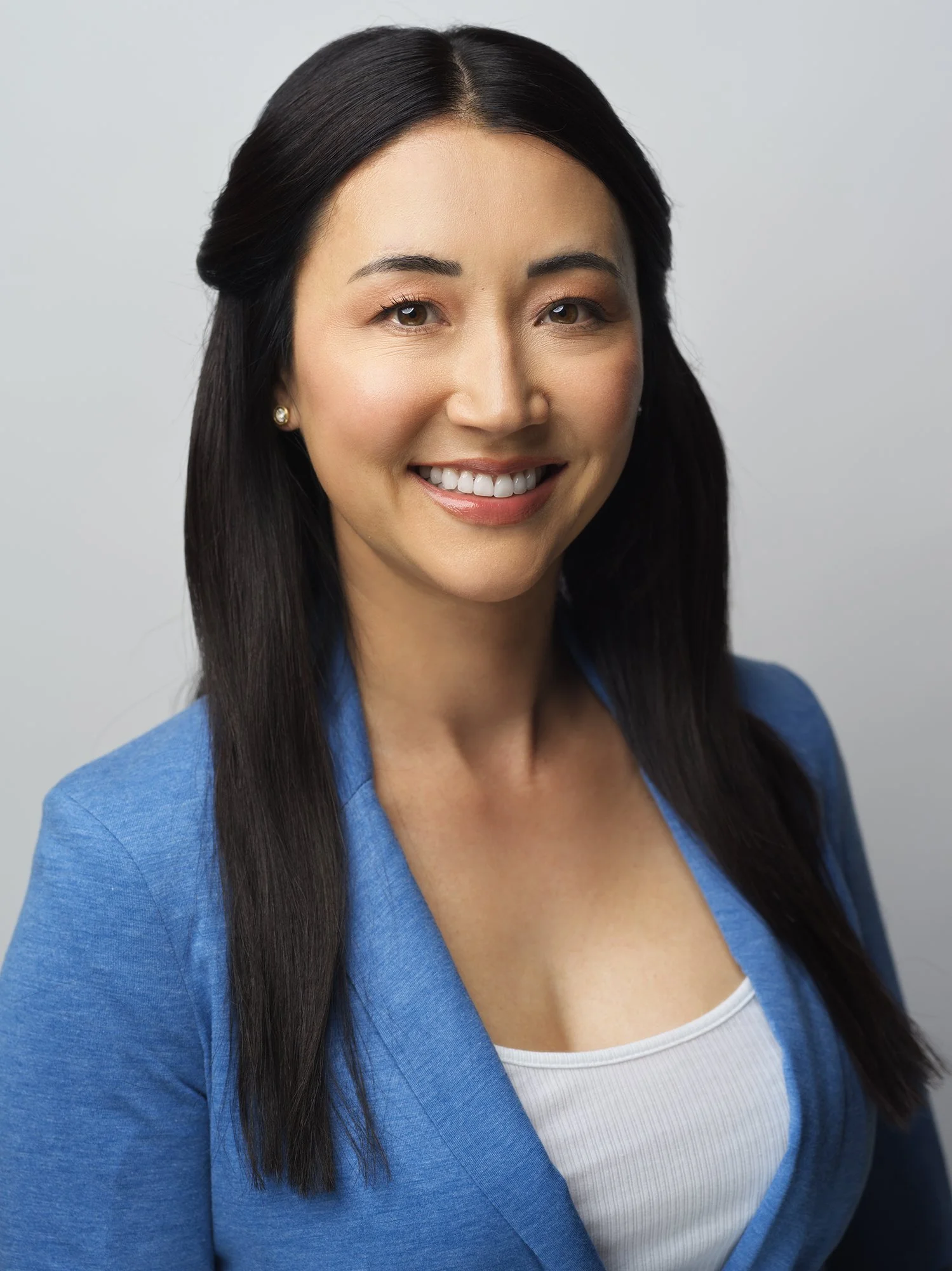 A woman with long dark hair, wearing a blue blazer and a white top, smiling against a plain gray background.