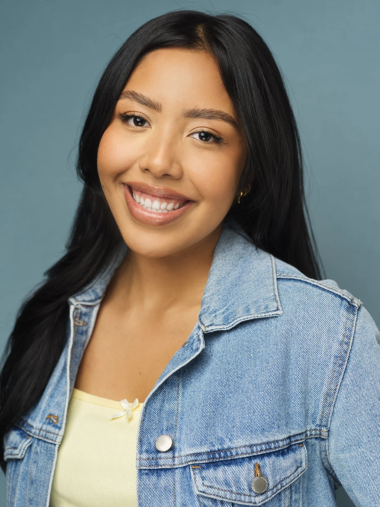 Close-up portrait of a smiling young woman with long black hair, wearing a denim jacket over a yellow top, against a blue background.