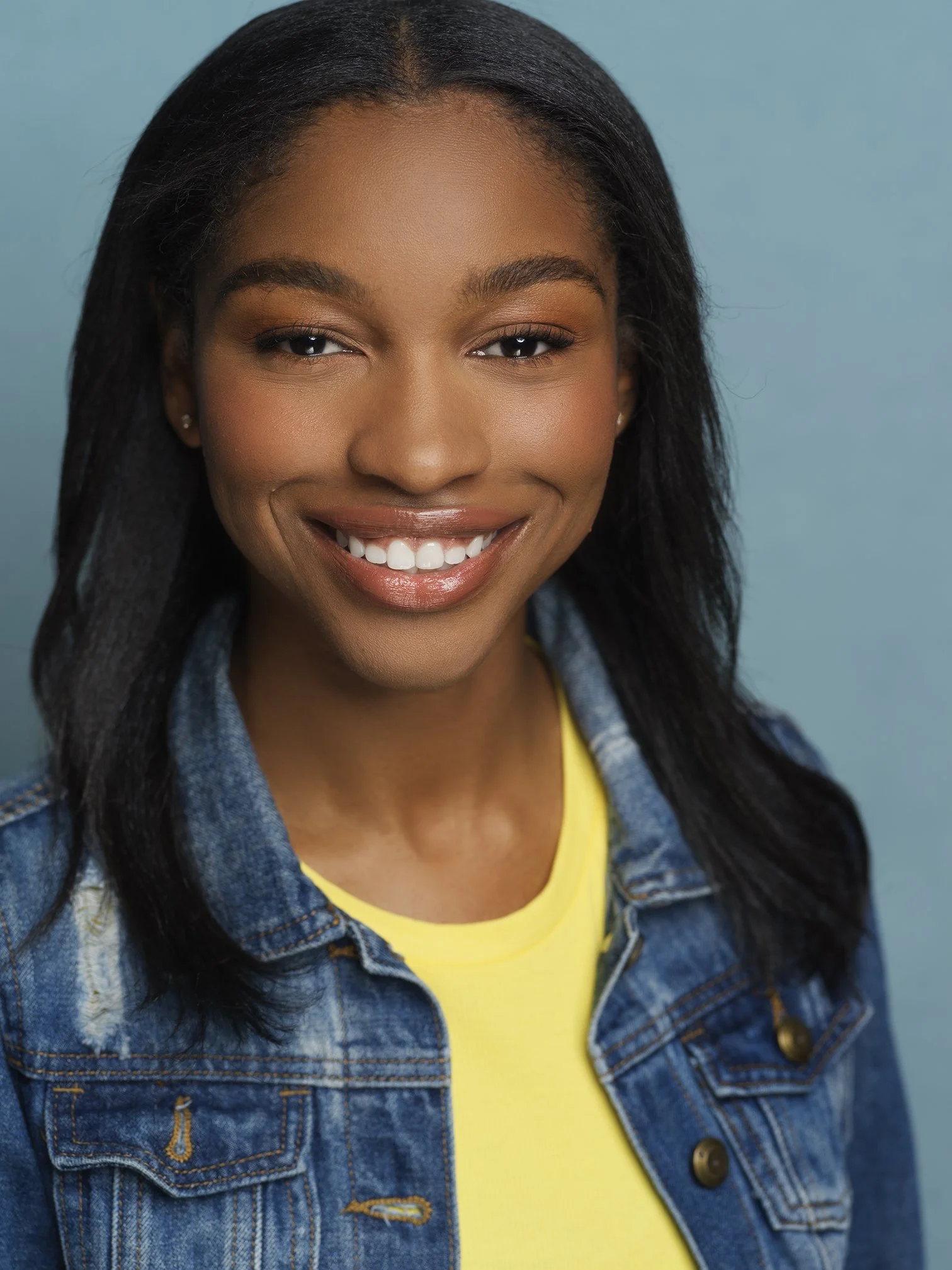 A young woman with dark hair, smiling and wearing a yellow shirt and denim jacket against a light blue background.