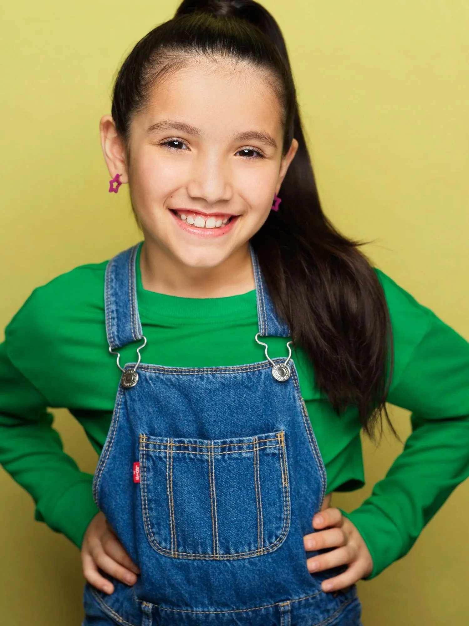 A young girl with long dark hair in a high ponytail, wearing a green long-sleeve shirt, denim overalls, pink star-shaped earrings, and smiling confidently against a yellow background.