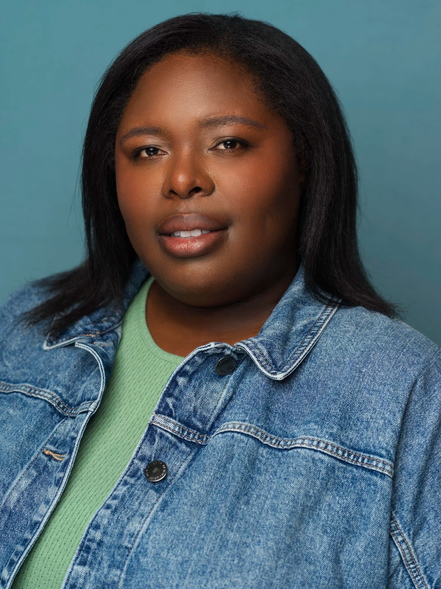 Close-up portrait of a woman with dark hair and brown skin, wearing a denim jacket over a green top, against a plain light blue background.