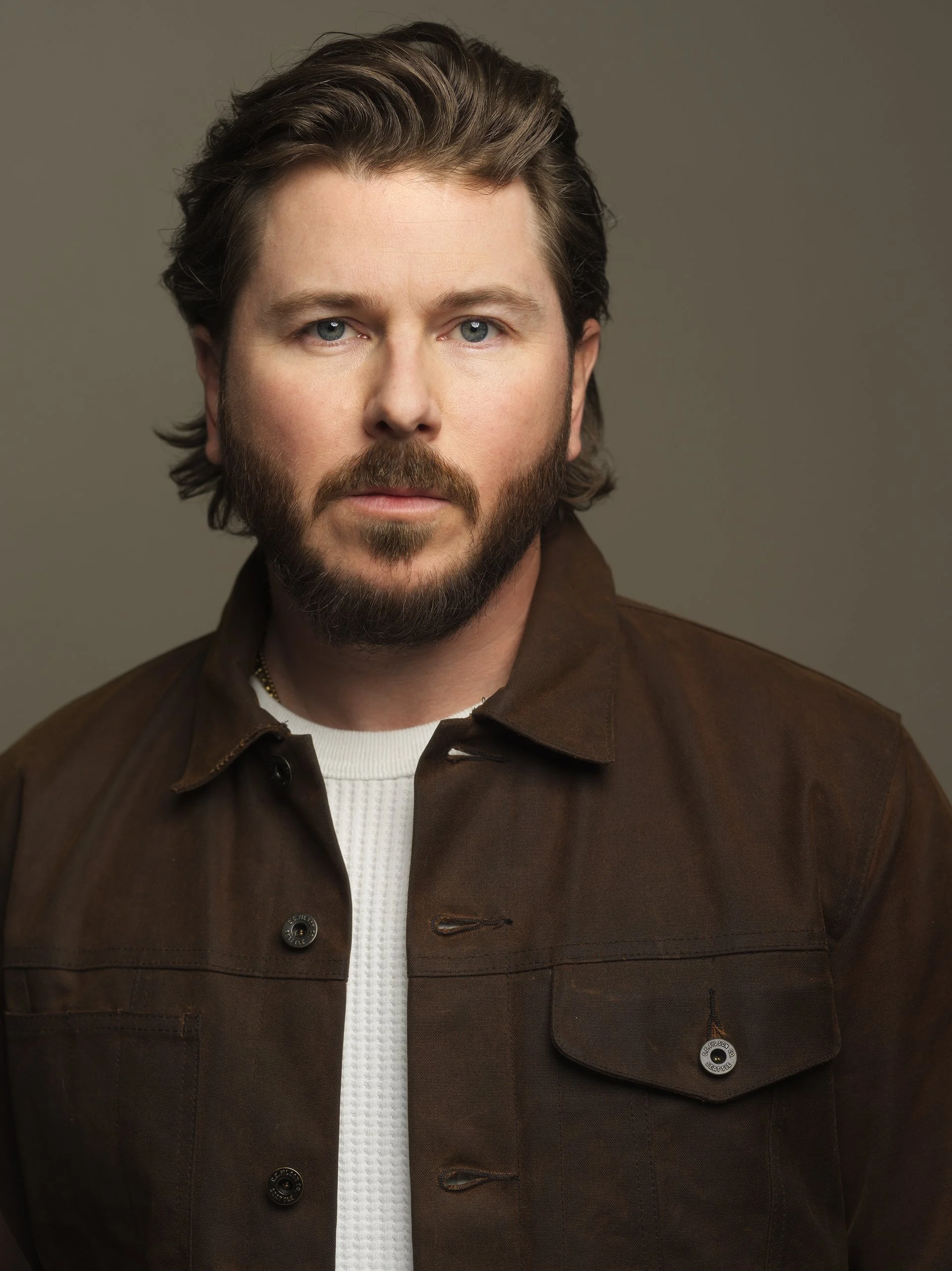 A close-up portrait of a man with brown hair, blue eyes, and a beard, wearing a brown jacket over a white shirt, against a neutral background.