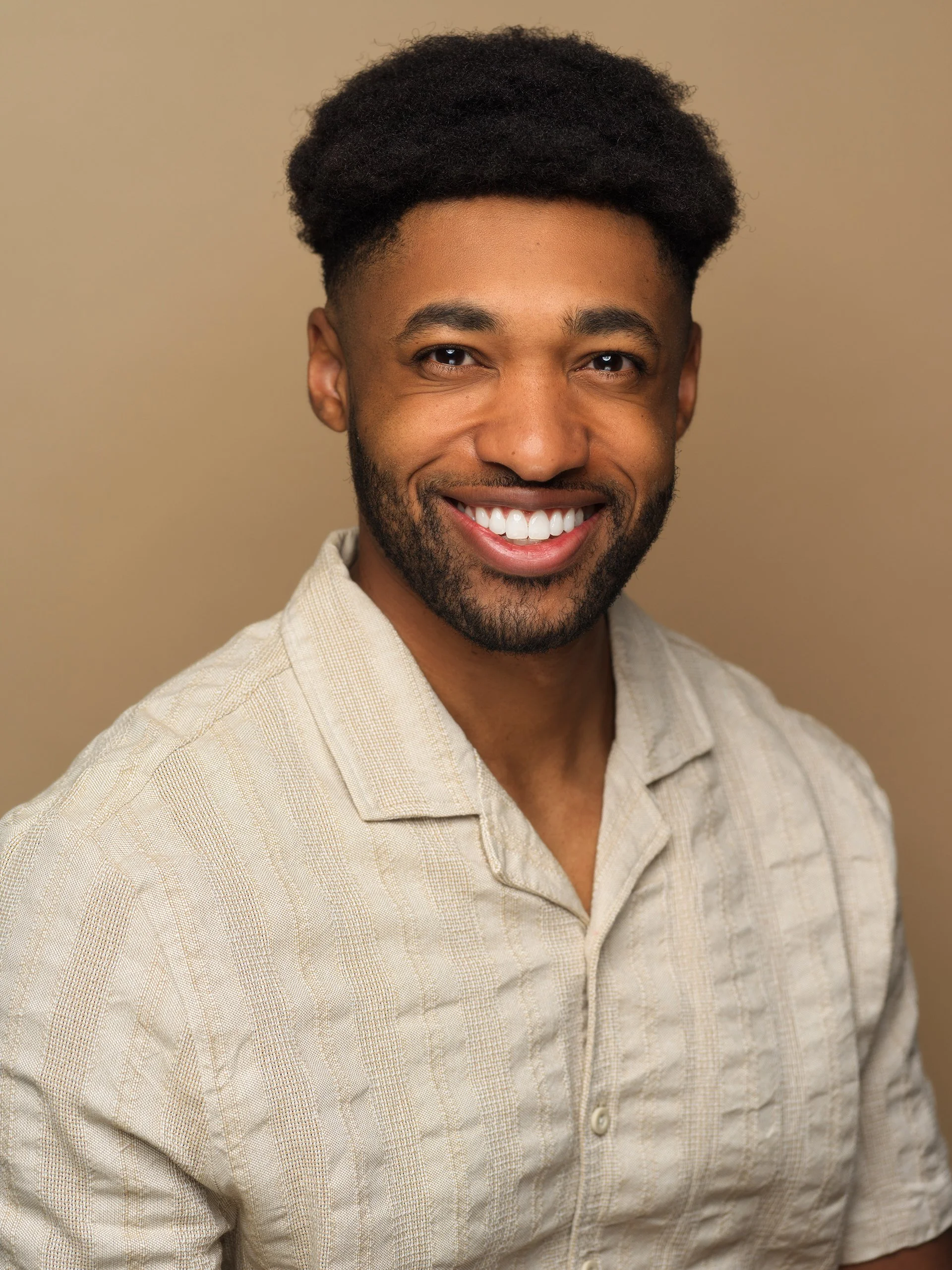 A young man with dark hair and a beard, smiling and wearing a beige button-up shirt, standing against a beige background.