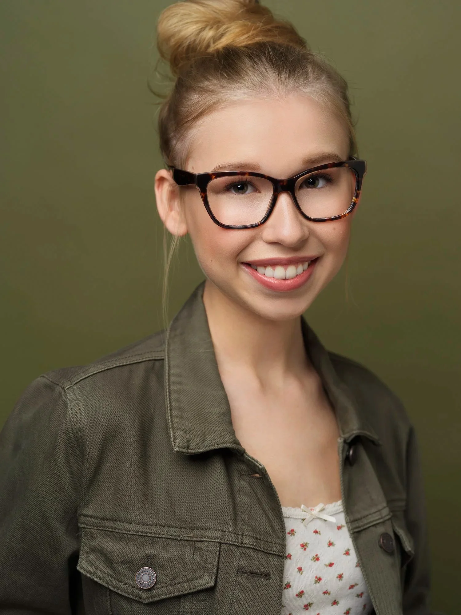 A young woman with blonde hair styled in a bun, wearing tortoise shell glasses, a dark green jacket, and a floral top, smiling against a green background.