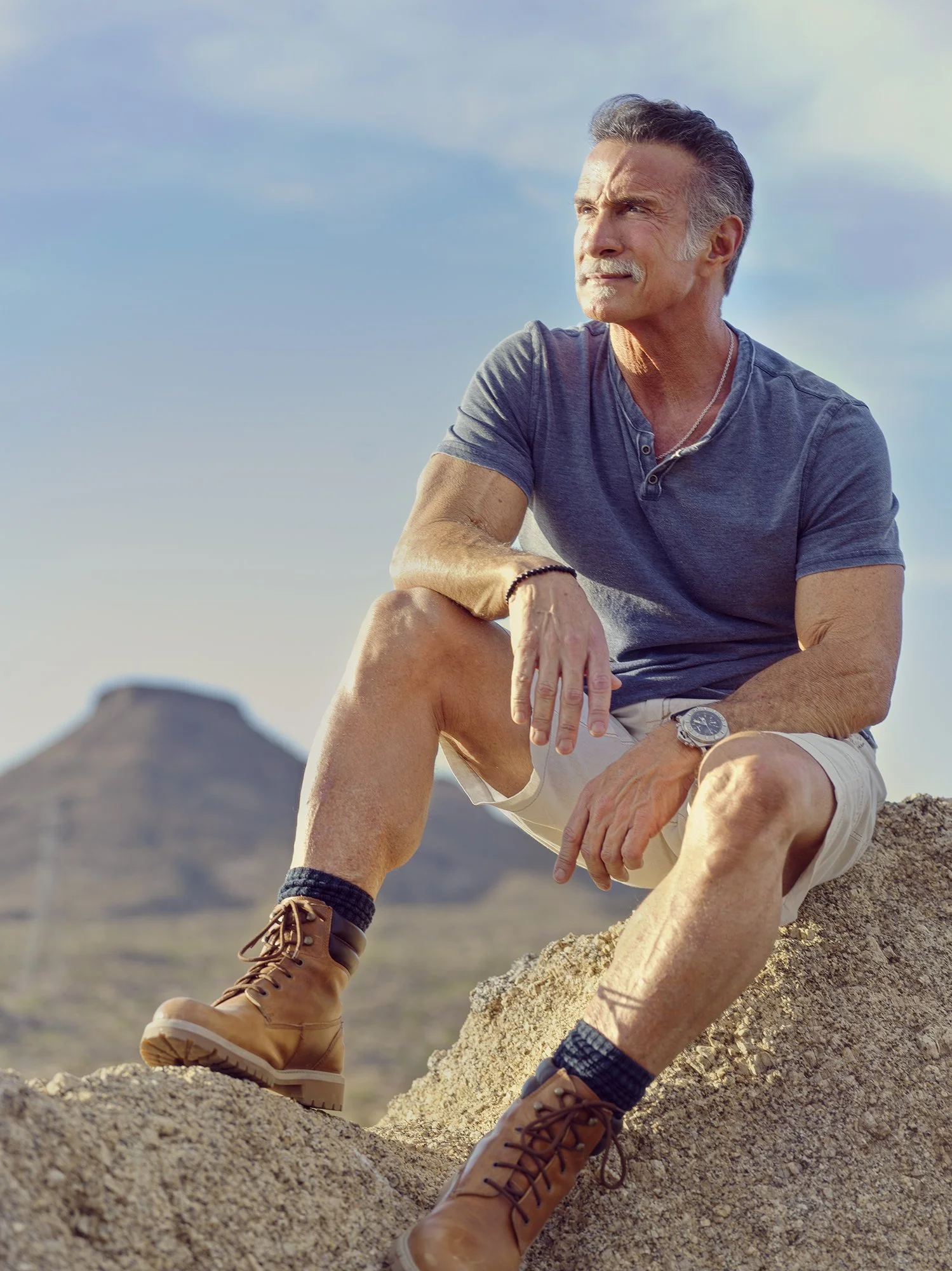 An older man sitting on a rock in a desert landscape, wearing a blue t-shirt, beige shorts, and tan boots, looking thoughtfully into the distance against a blue sky.