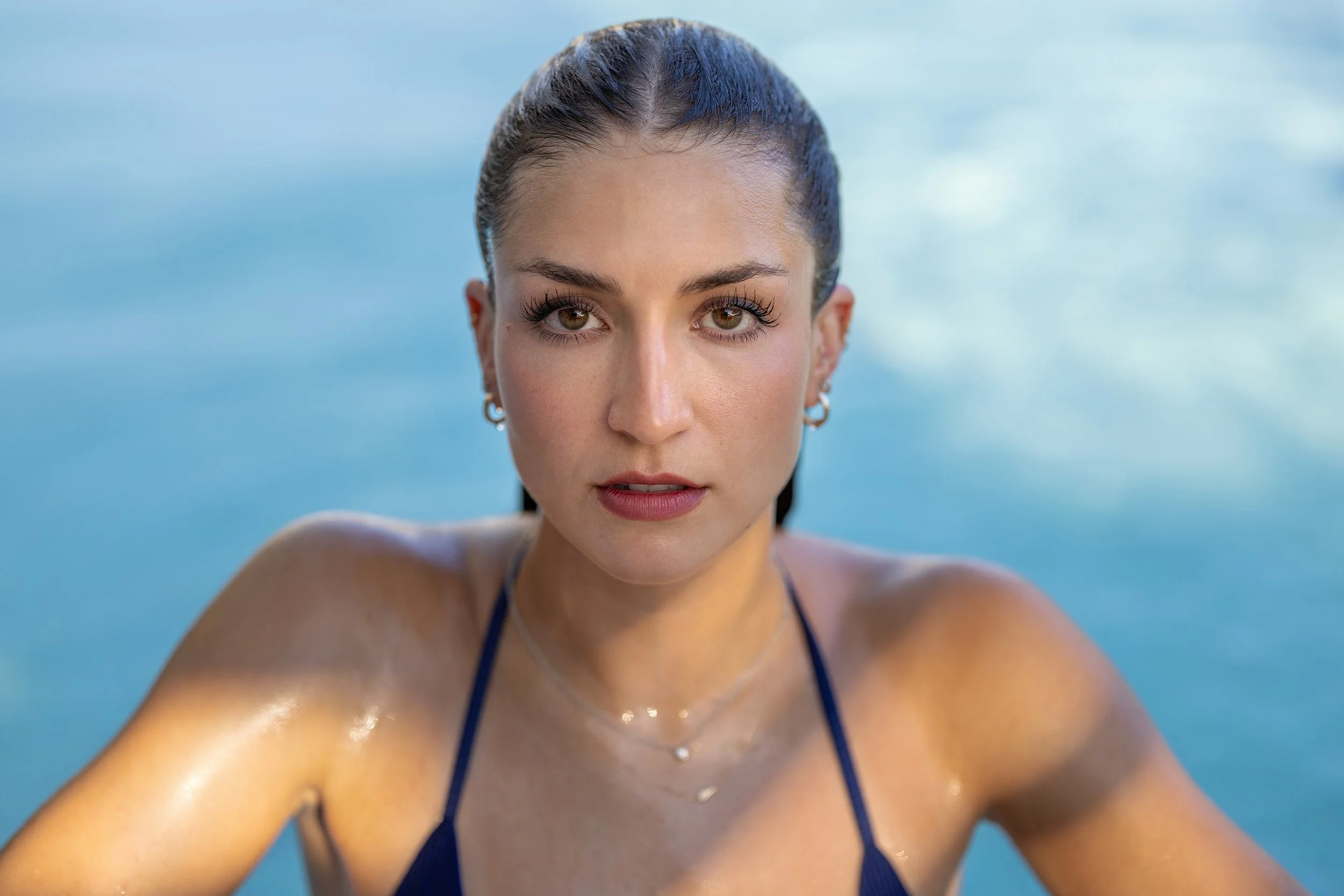 Close-up of a woman with wet hair, wearing earrings and a dark blue swimsuit, taking a selfie by a pool with a blue water background.