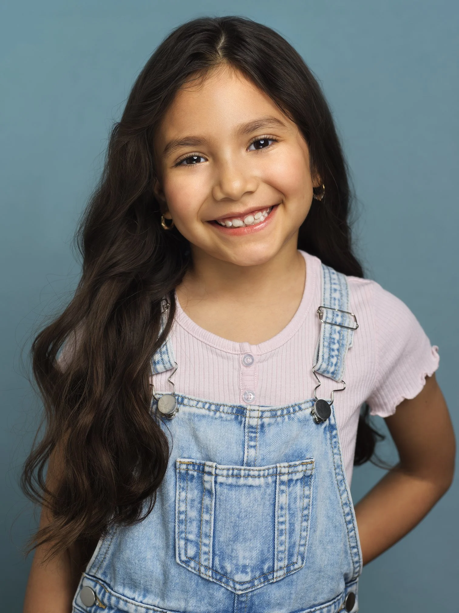 Close-up portrait of a young girl with long wavy dark hair, smiling, wearing hoop earrings, a pink shirt with buttons, and denim overalls against a blue background.