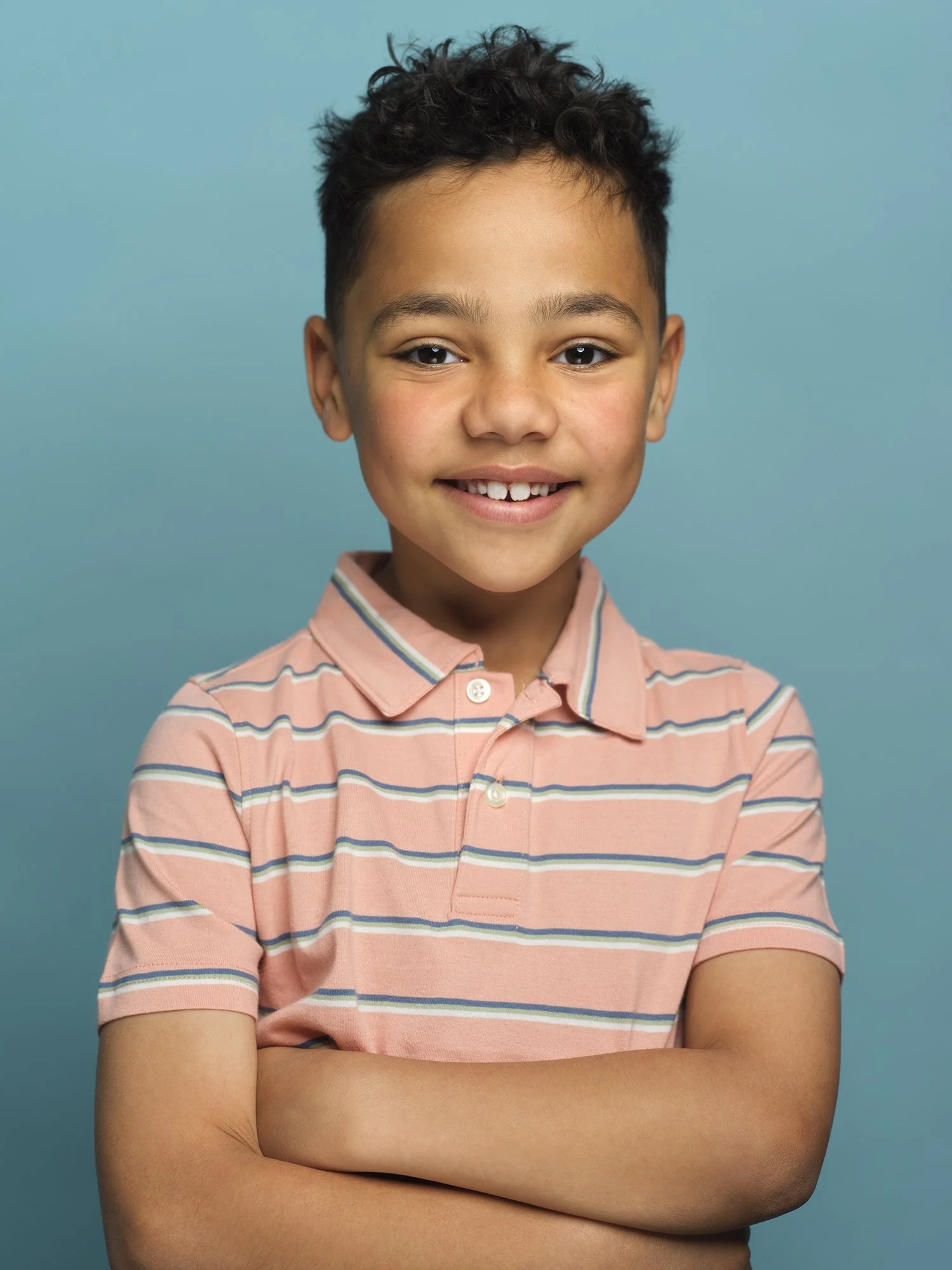 Portrait of a young boy with curly dark hair, wearing a pink and striped polo shirt, smiling with crossed arms and standing against a blue background.