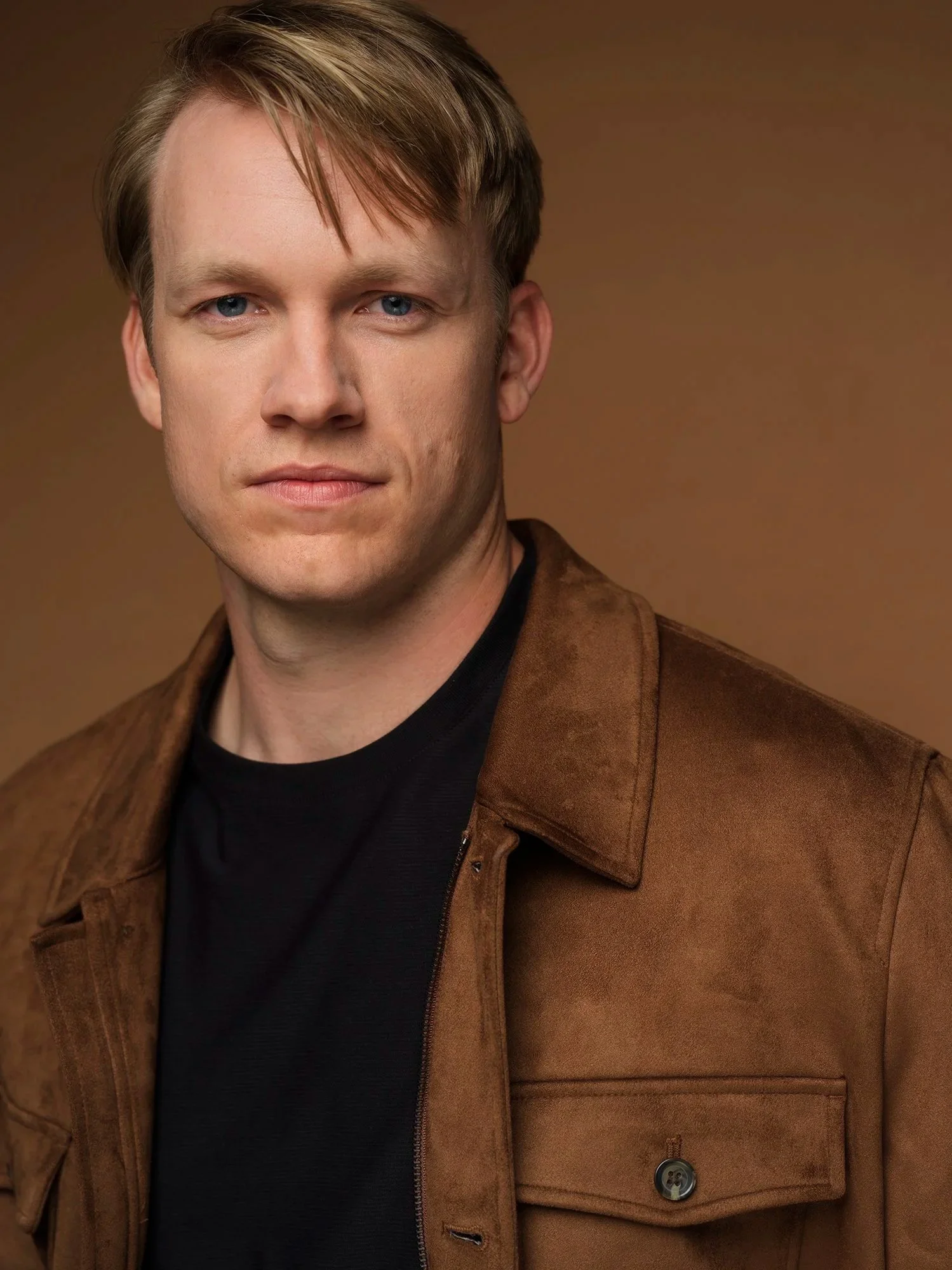 Portrait of a man with light skin, blue eyes, and brown hair, wearing a brown jacket and black shirt, against a brown background.