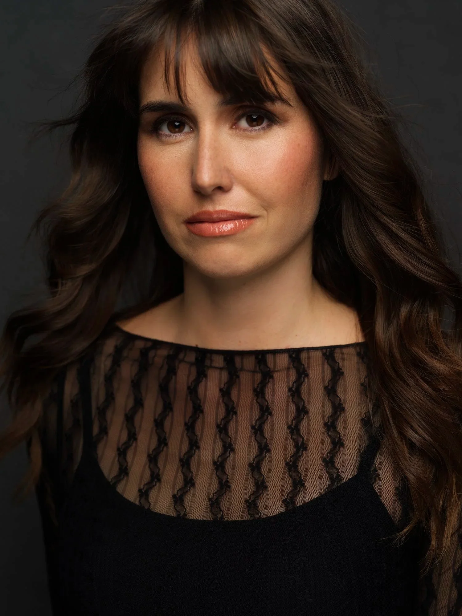 Close-up of a woman's face with brown hair, wearing a black top with a sheer lace pattern around the neckline, against a dark background.