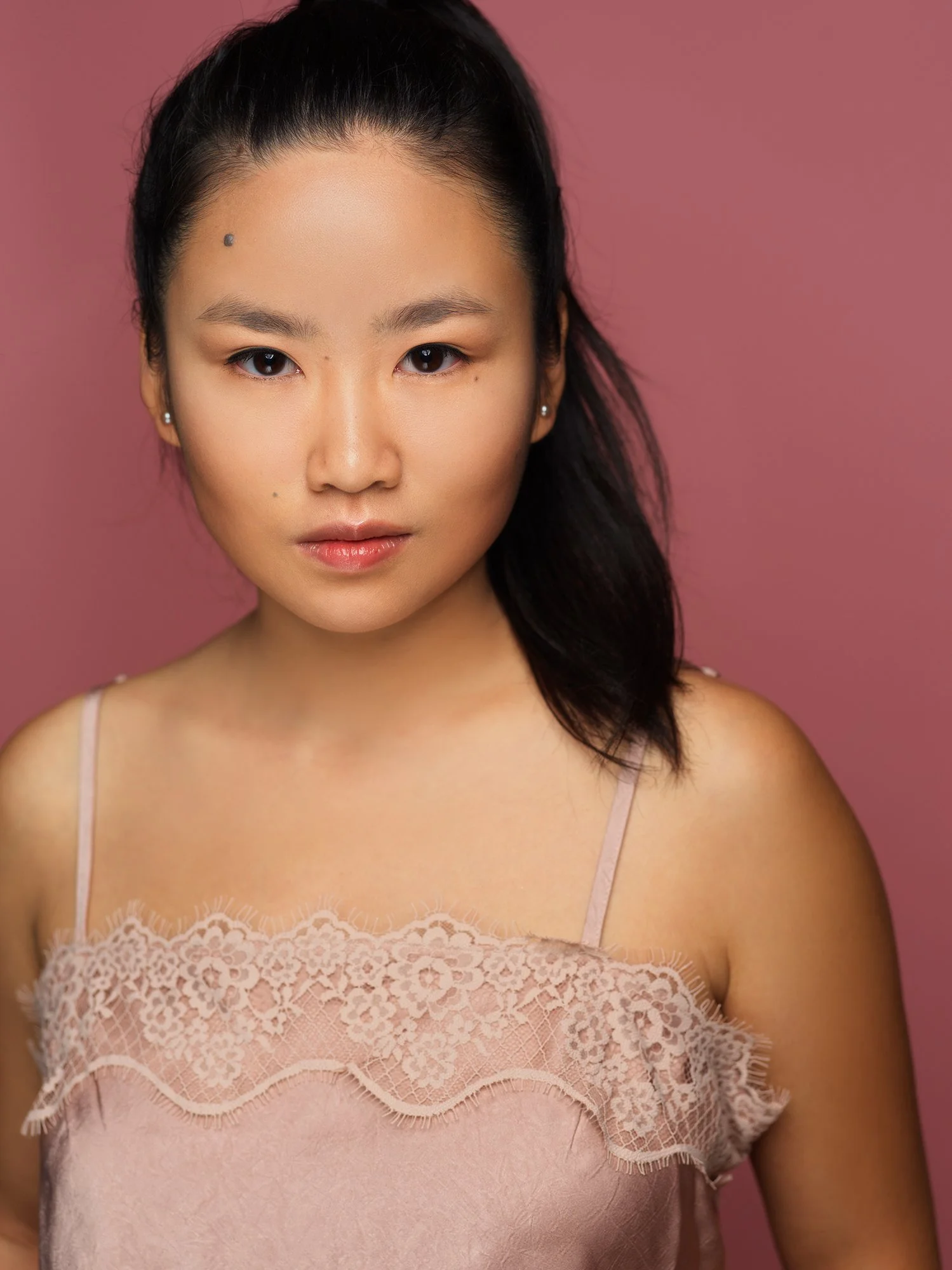 Close-up of a woman with dark hair, wearing a light pink lace top, standing against a pink background.