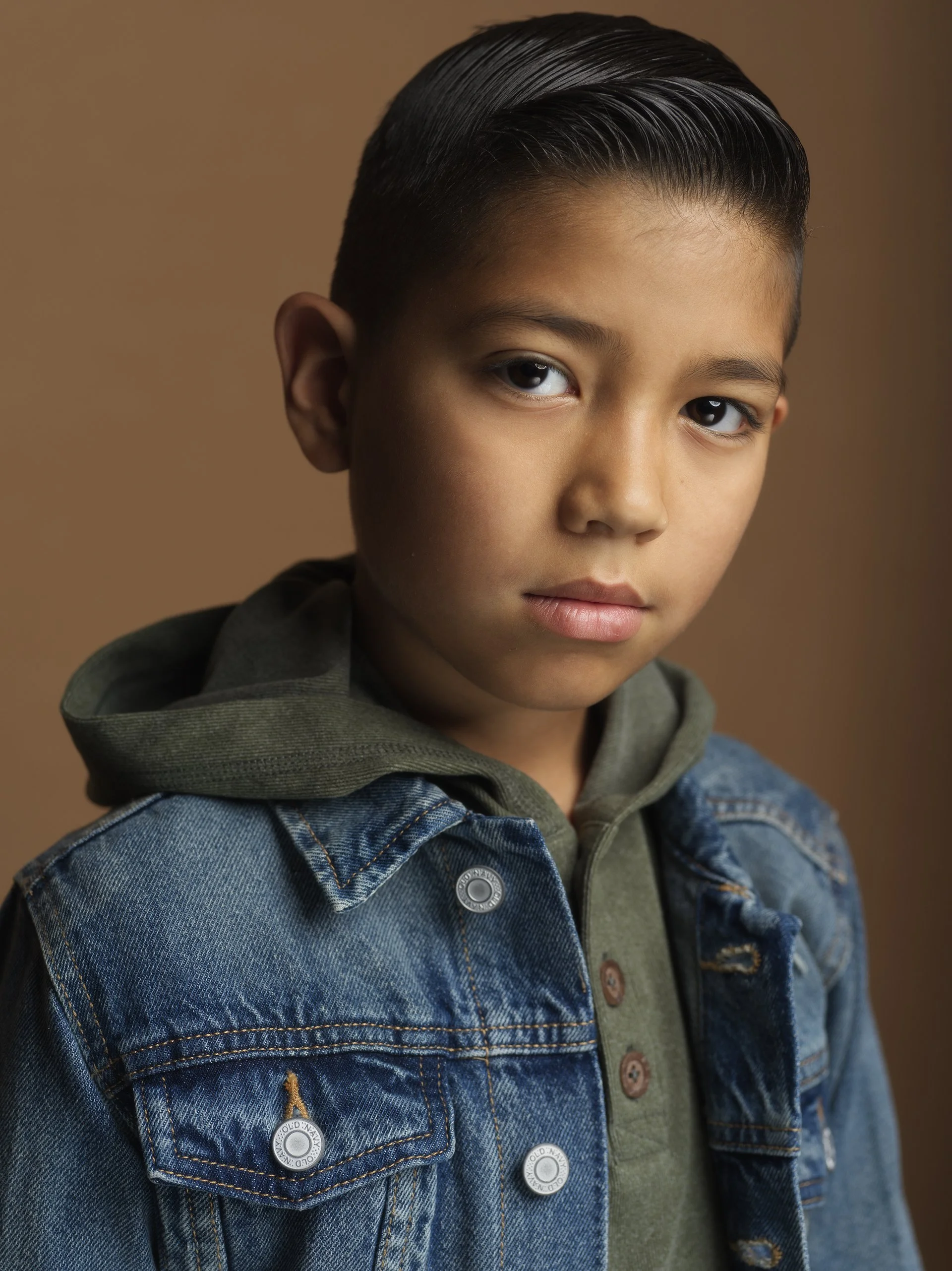 Close-up of a young boy with short, slicked-back hair, wearing a denim jacket over a green hoodie, against a brown background.