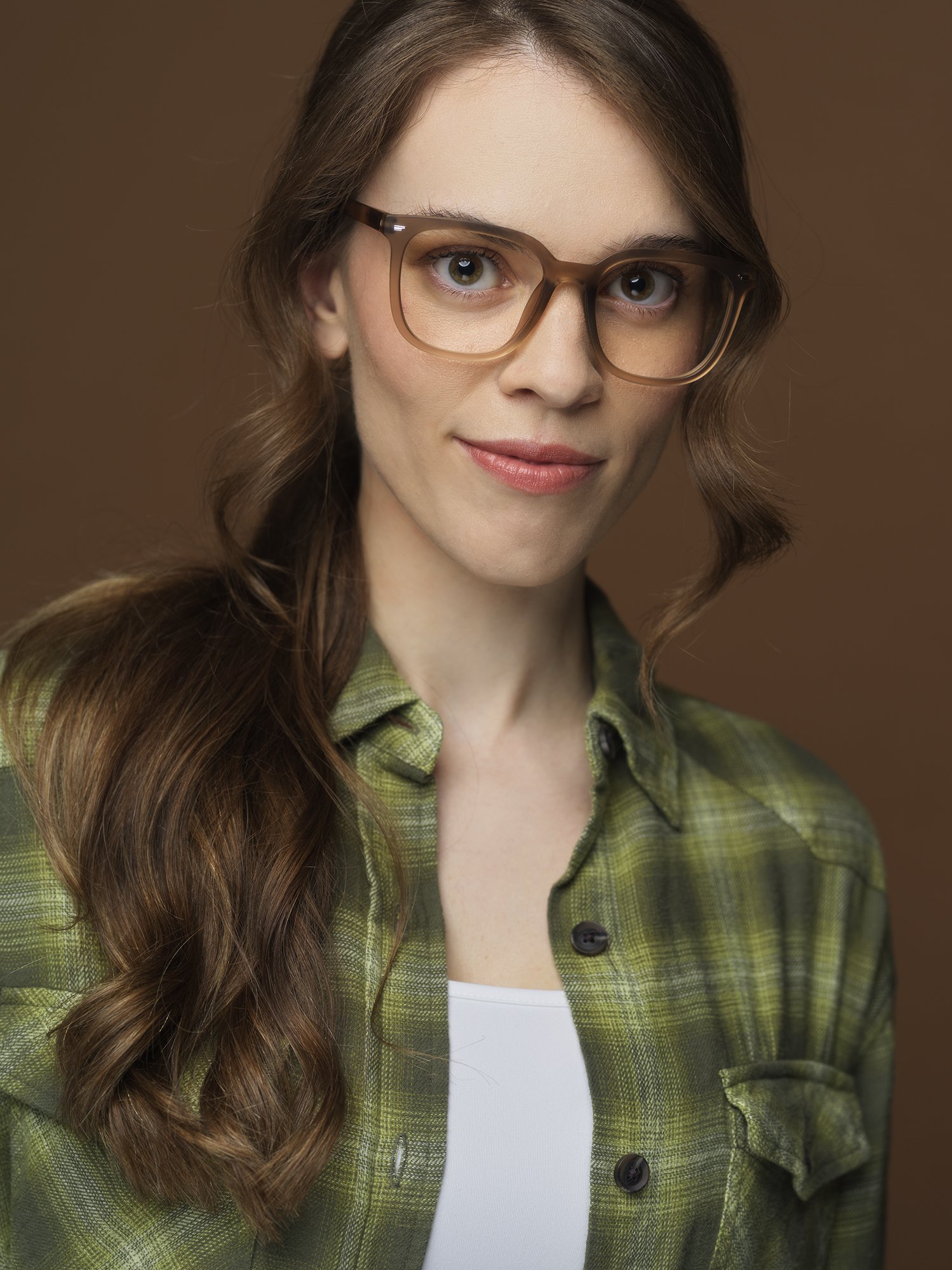 Close-up of a young woman with long, wavy auburn hair, wearing large, brown glasses, a green plaid shirt, and a white top, smiling softly with a brown background.