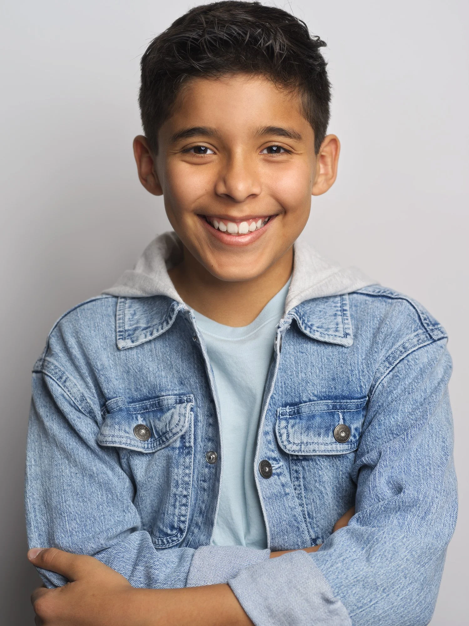 A young boy with dark hair smiling, wearing a light blue shirt and a denim jacket with buttons, standing against a plain grey background.