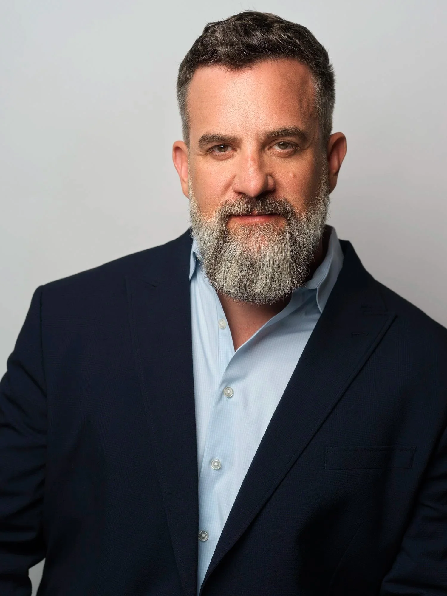 A middle-aged man with dark hair, a salt-and-pepper beard, wearing a navy suit and a light blue shirt, looking into the camera with a serious expression.