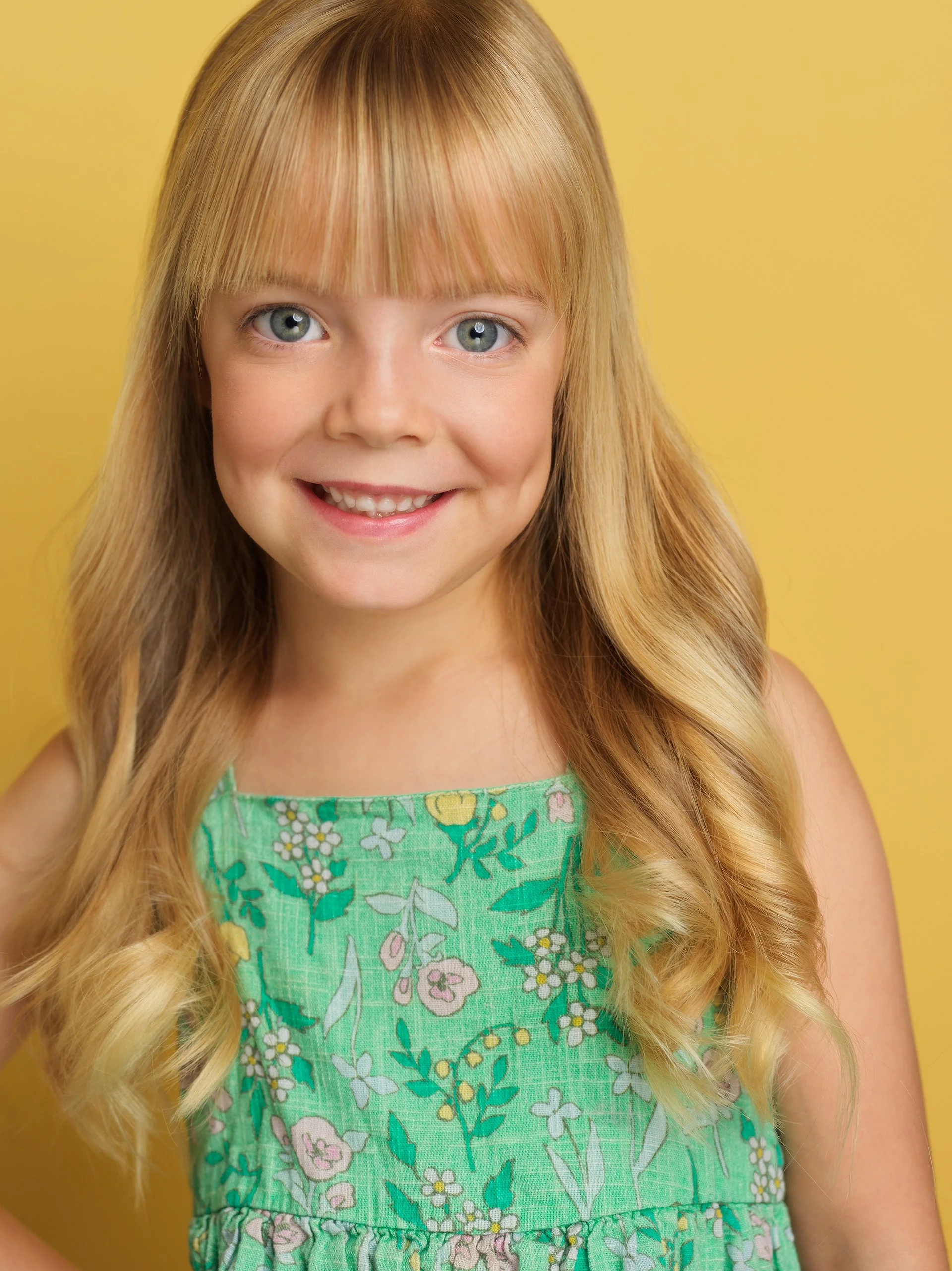 Close-up of a young girl with long wavy blonde hair, blue eyes, a big smile, wearing a green floral dress, against a yellow background.