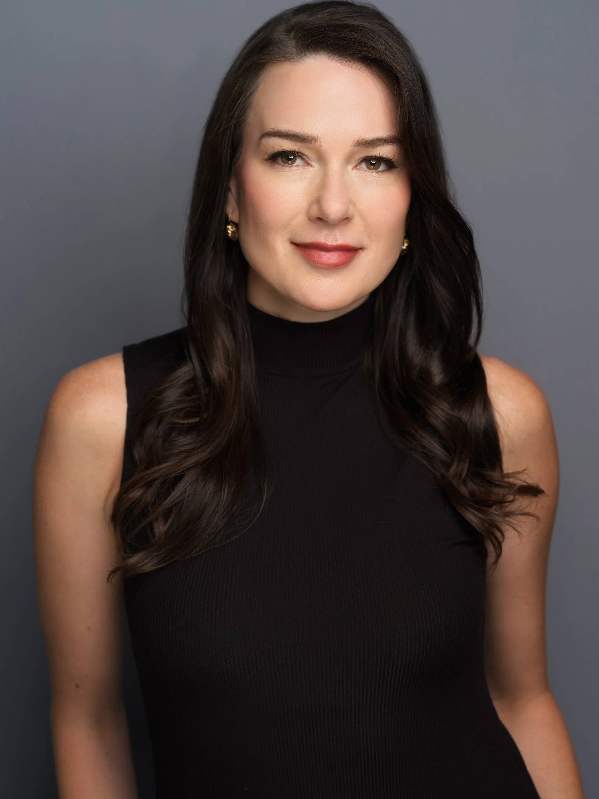 Close-up portrait of a woman with long brown wavy hair, wearing a black sleeveless top and gold earrings, against a gray background.