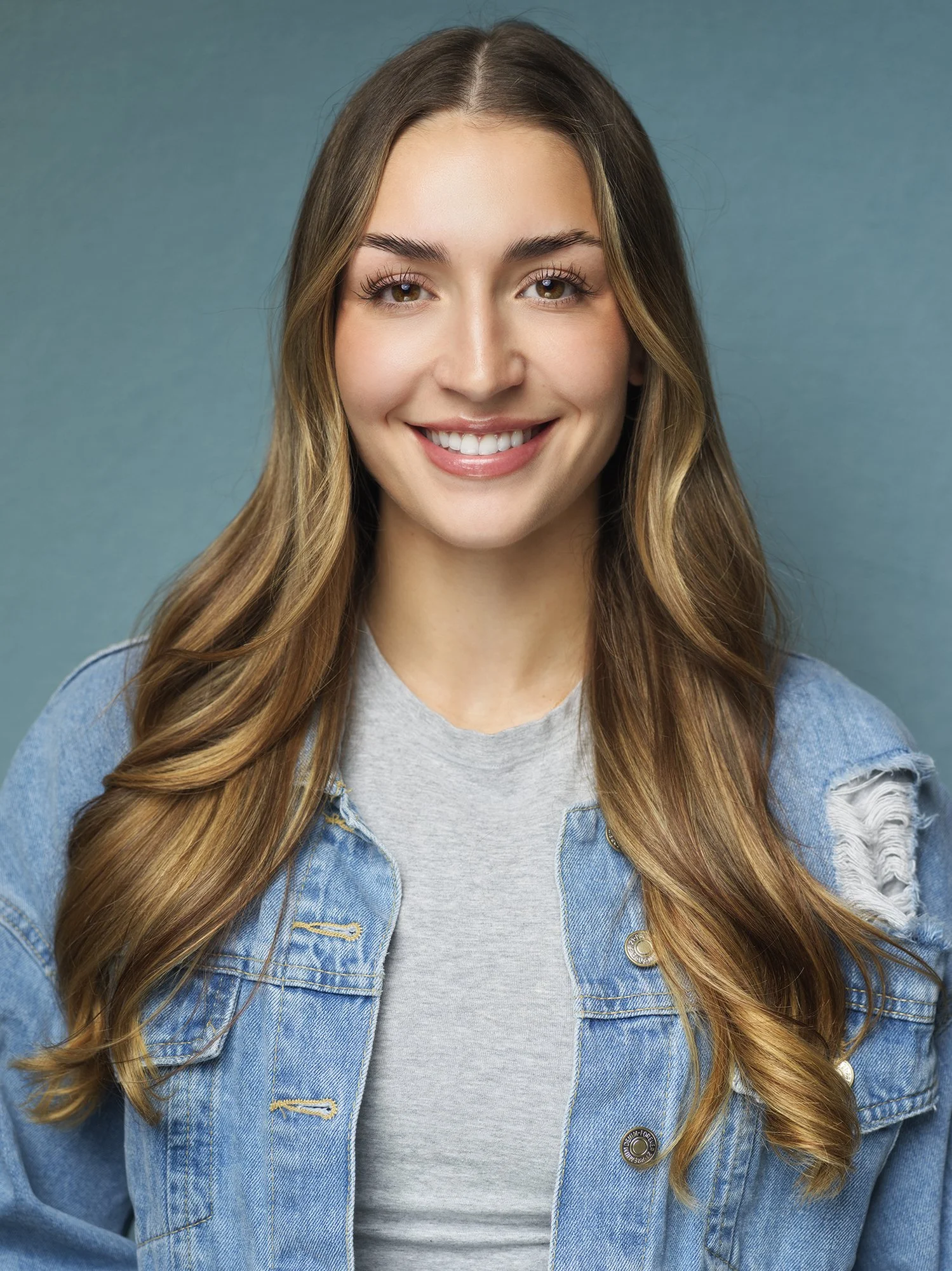 Close-up of a young woman smiling, with long wavy hair, wearing a light gray T-shirt and a blue denim jacket, against a teal background.