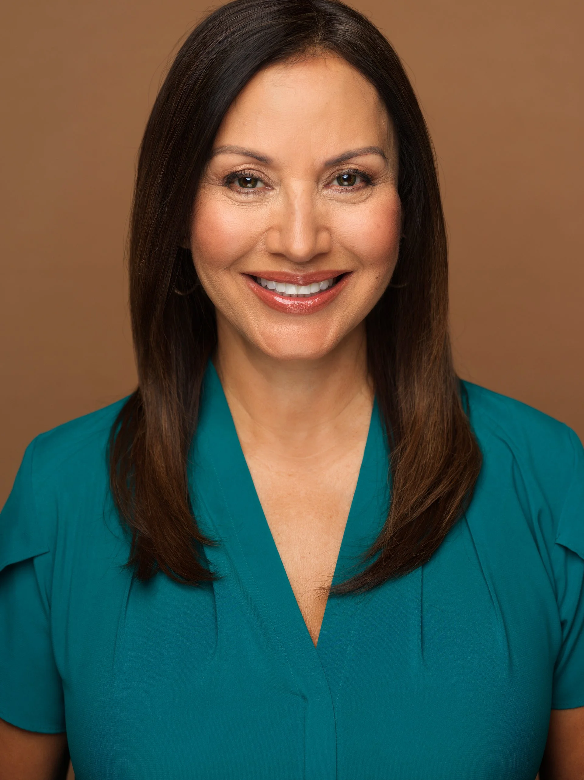 Close-up of a woman with dark hair smiling in a teal top against a brown background.