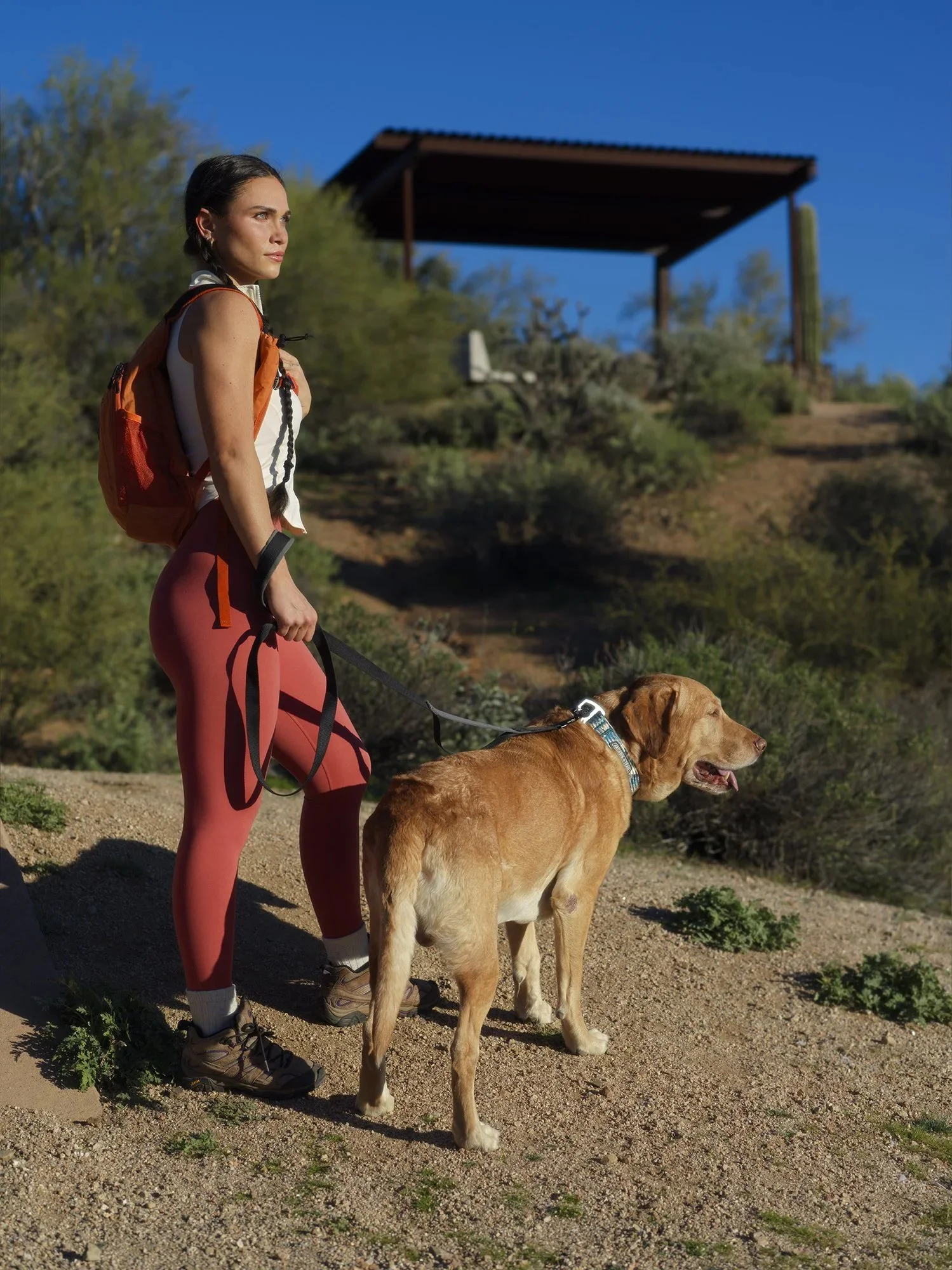 A woman with dark hair tied back, wearing a white sleeveless top, red leggings, hiking boots, and carrying an orange backpack, walking her golden retriever dog on a dirt trail in a desert landscape with bushes, cactus, and a wooden structure in the b