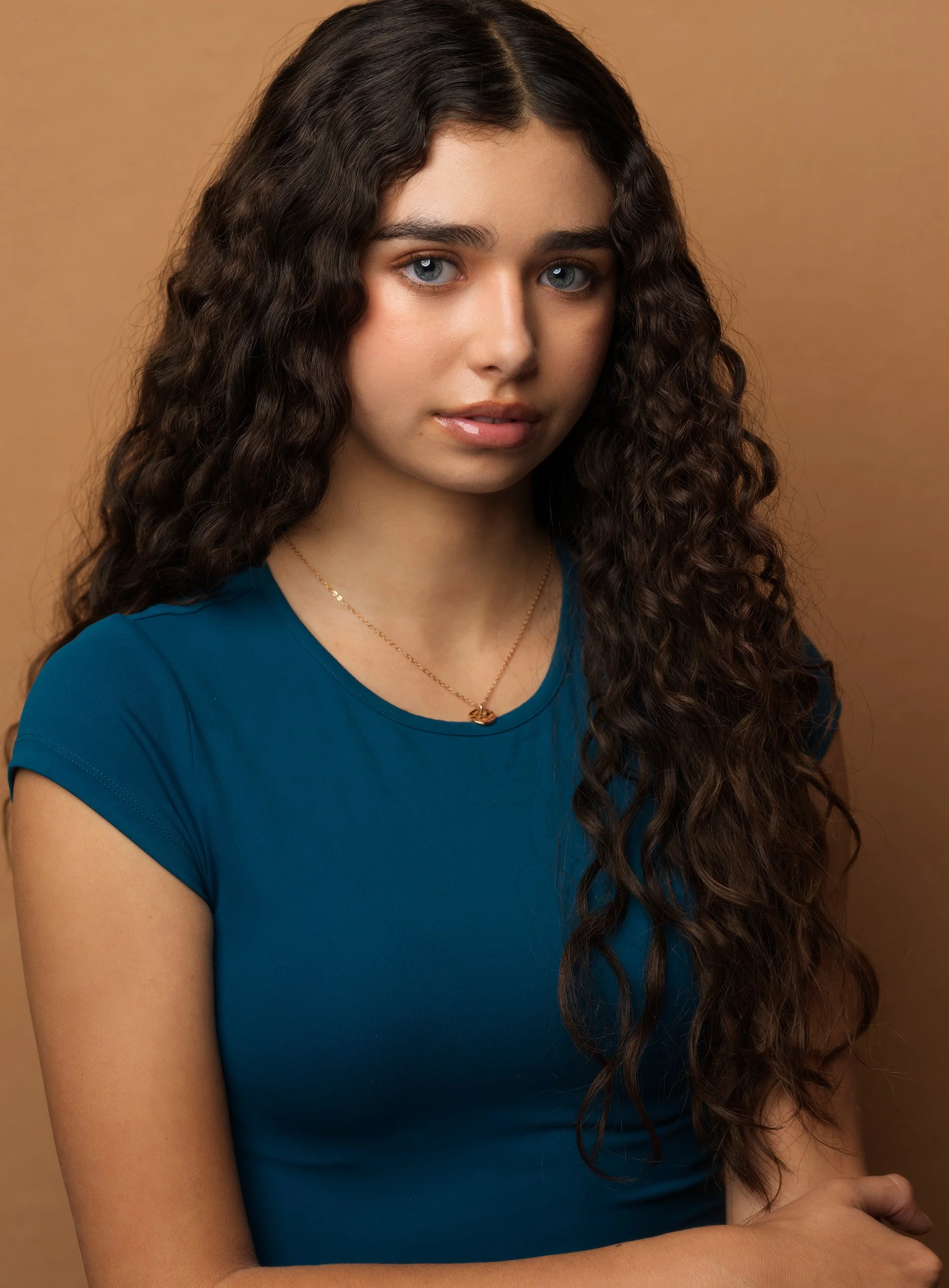 A young woman with long, curly brown hair, blue eyes, and light skin, wearing a blue top and a delicate gold necklace, standing against a plain brown background.