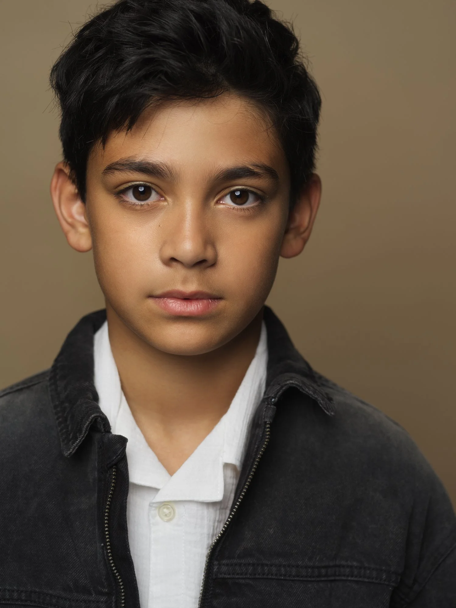 Close-up portrait of a young boy with dark hair, brown eyes, and a neutral expression, wearing a white collared shirt and a dark zip-up jacket, against a plain beige background.