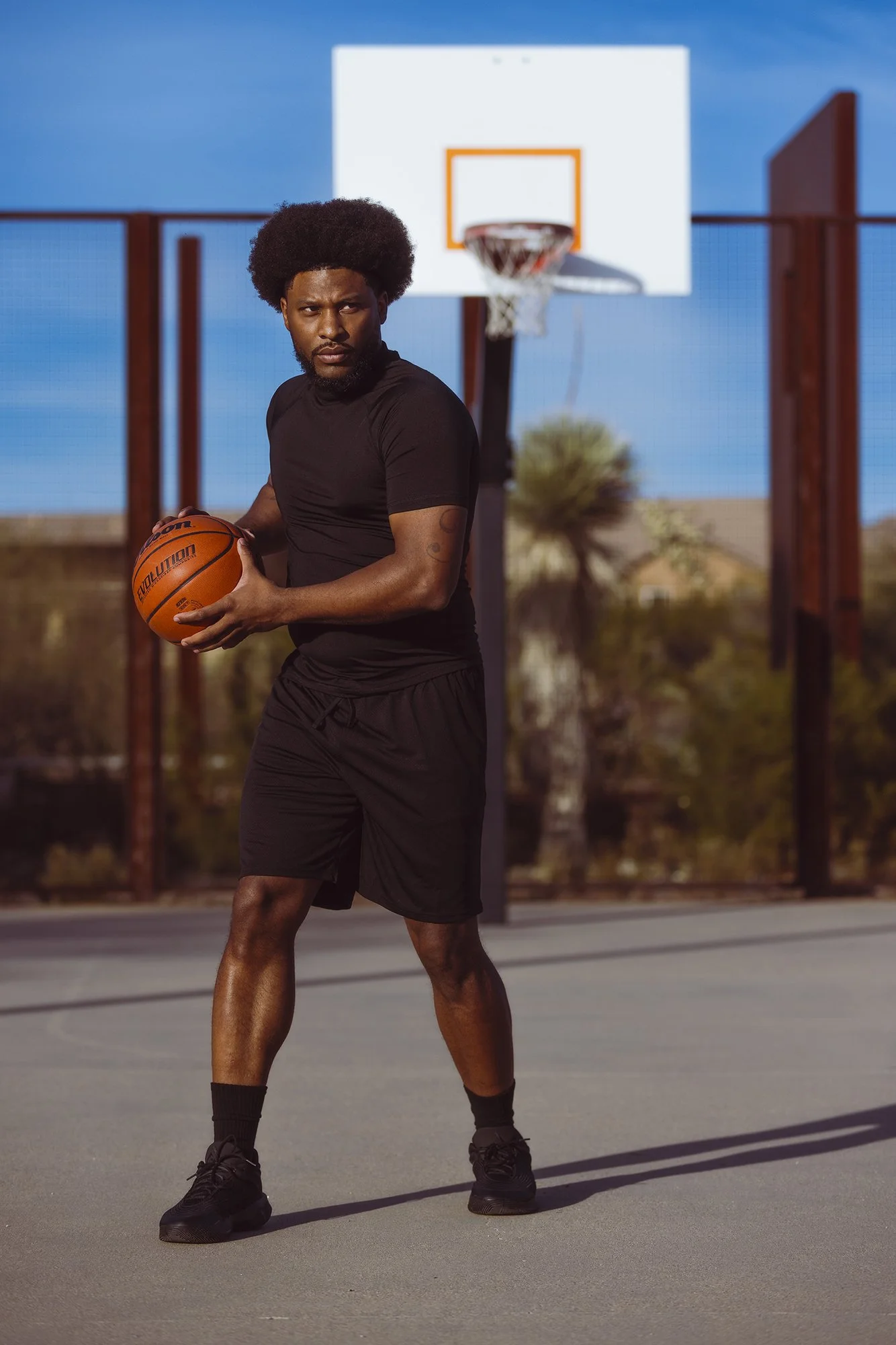A man with an afro hairstyle and beard standing on an outdoor basketball court, dressed in black athletic clothing, holding a basketball, with a basketball hoop in the background under a blue sky.