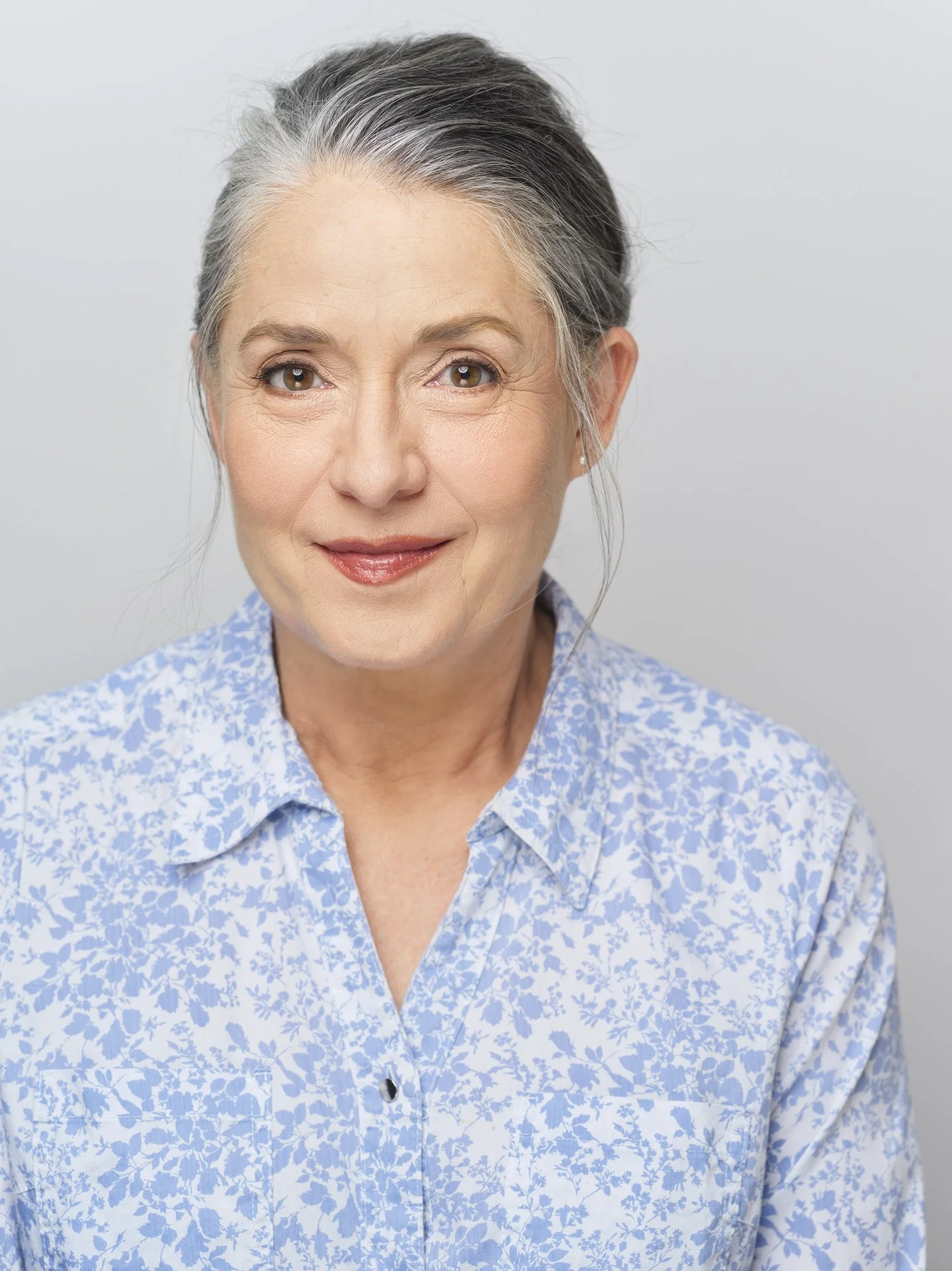 Close-up of an older woman with gray hair, smiling, wearing a light blue floral blouse, standing against a light gray background.