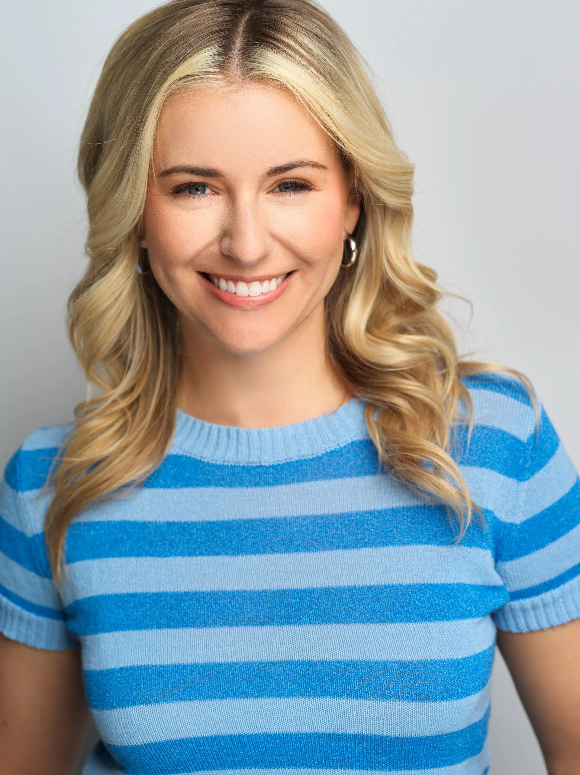 A woman with long blonde wavy hair wearing a blue and white striped short-sleeve sweater, smiling, with a plain light gray background.