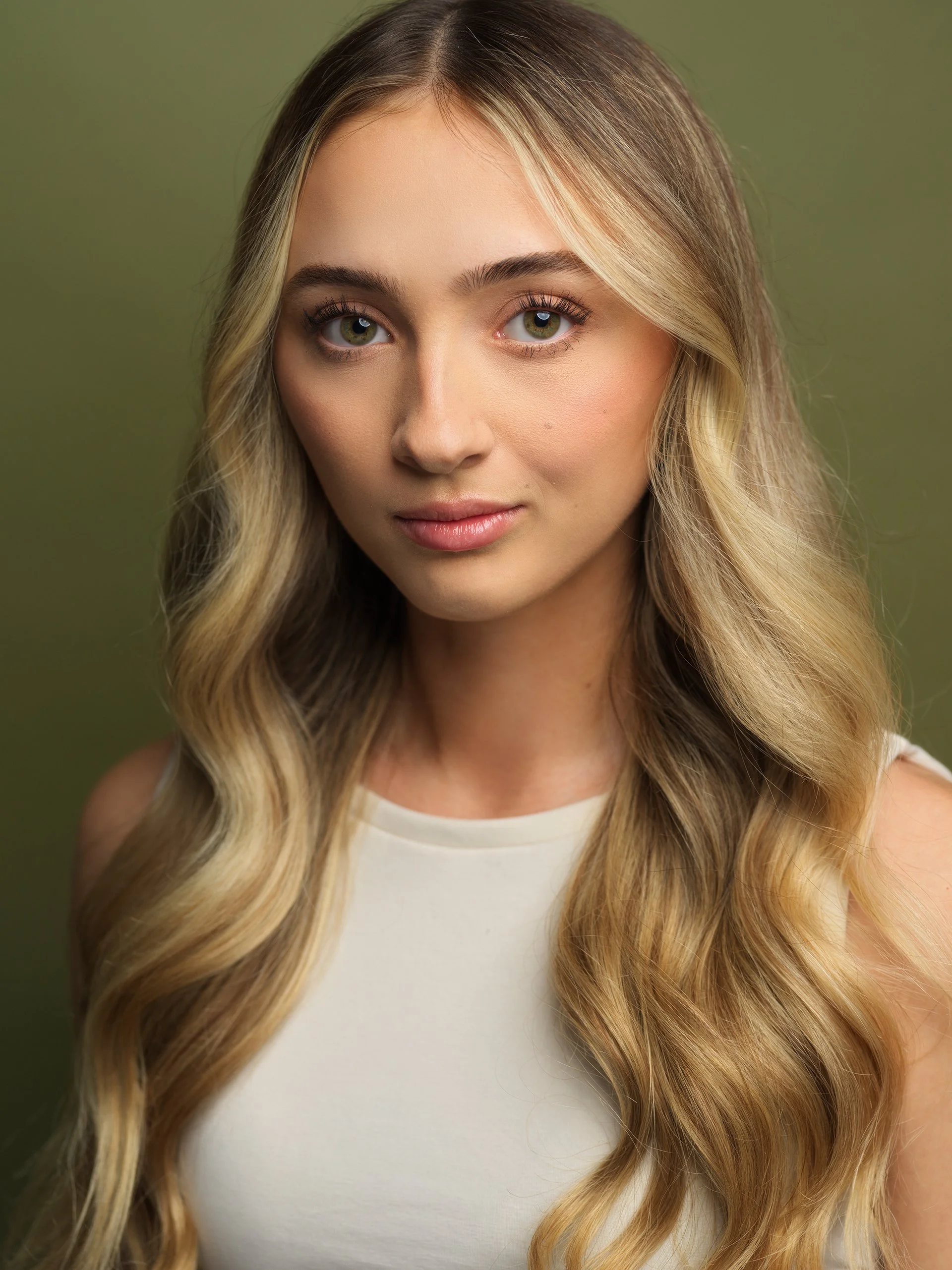Close-up of a young woman with long, blonde wavy hair and light green eyes, wearing a white sleeveless top, against a green background.