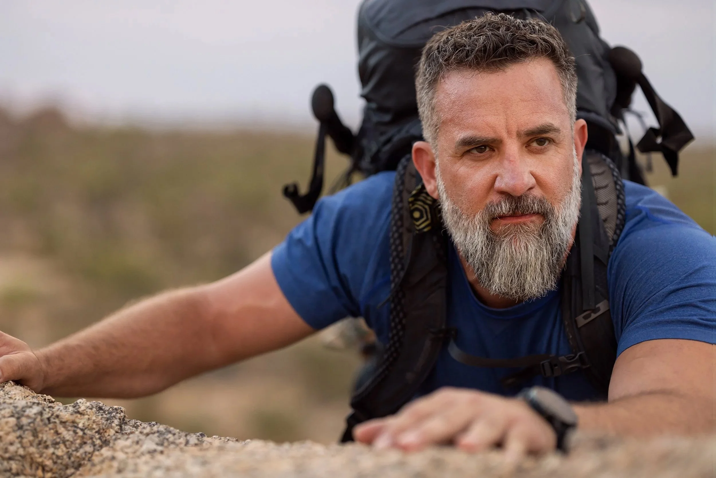 A man with a gray beard and short, curly gray hair crawling on the ground outdoors while carrying a large black backpack.